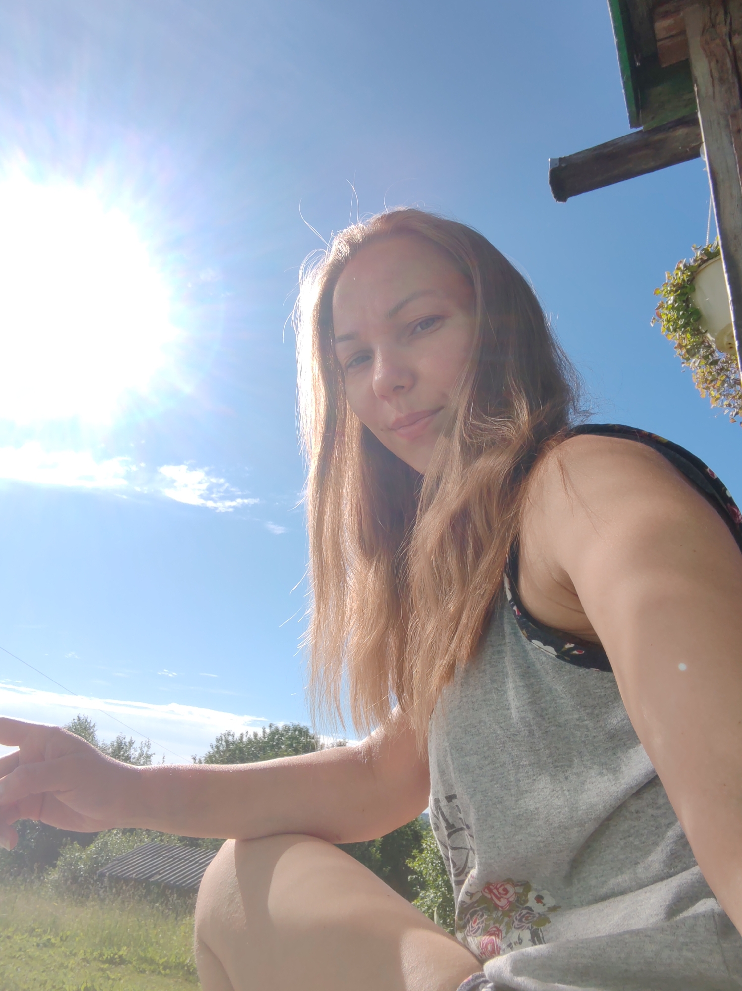 A young woman with light brown hair is captured in a selfie outdoors. She is wearing a grey tank top with a floral pattern visible on the straps and hem. The background consists of a clear blue sky with the sun prominently featured in the upper left corner, casting lens flare. To the right, part of a wooden structure with a green roof and hanging plants is visible. The lower portion of the image shows greenery, trees, and a small building with a corrugated roof, indicative of a rural or suburban setting in Чухлэм, Russia. The woman is looking towards the camera with a slight smile.