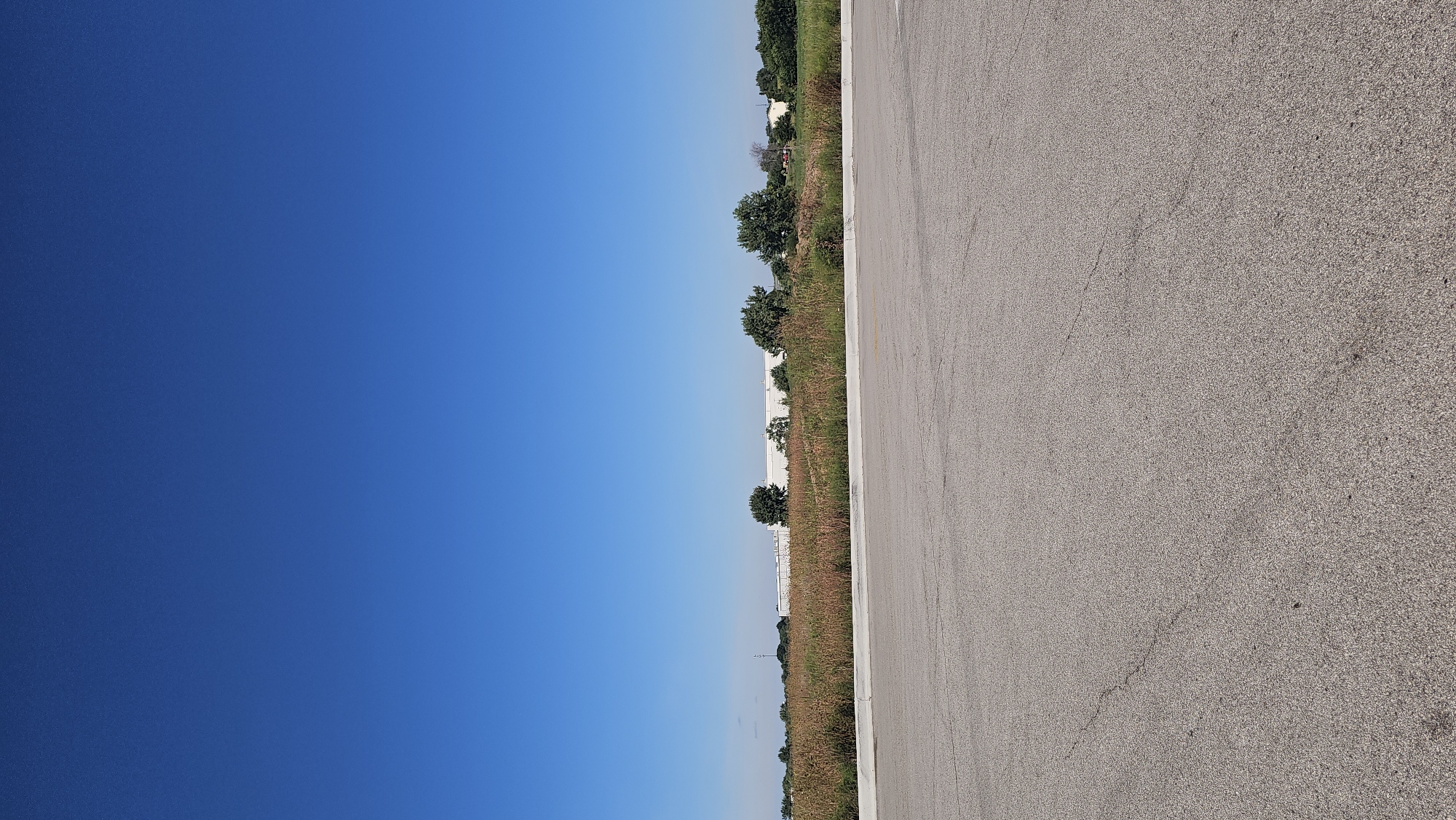 An aerial view captures a bright, cloudless blue sky dominating the upper portion of the image. Below the sky, a flat, open field with tall, dry grasses stretches across the mid-ground. A line of green trees is visible in the distance, breaking the horizon. Behind the trees, a large, long, white building with a flat roof is partially obscured. The foreground is an asphalt surface, appearing to be a road or parking lot, with visible cracks and a lighter grey border along the bottom edge. The scene is located in the city of Belvidere, United States.