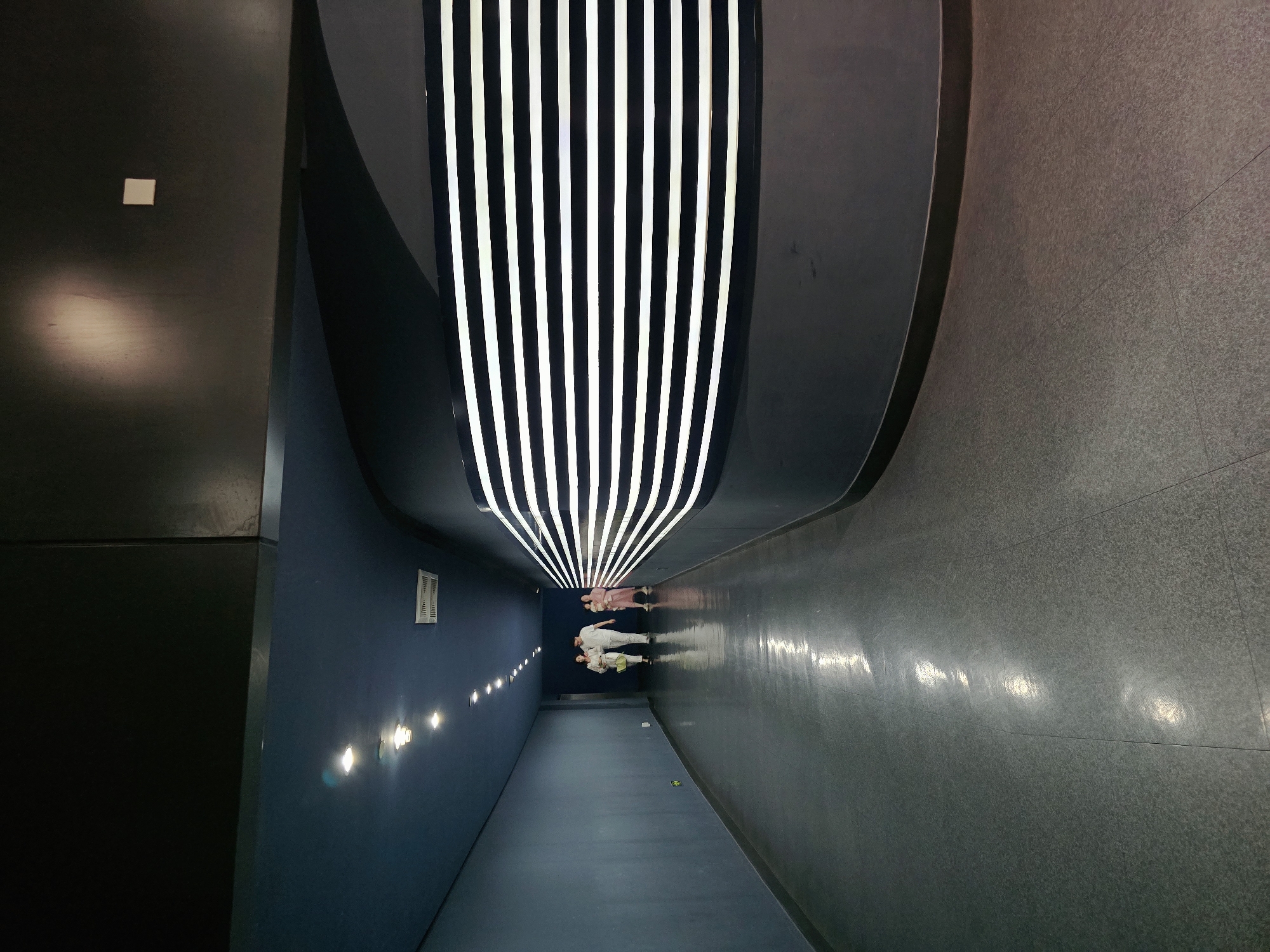 The image shows a long, dimly lit corridor with a blue ceiling and dark grey tiled floor. A series of spotlights are embedded in the ceiling, creating reflections on the floor. On the right side of the corridor, there is a curved dark wall with a decorative panel of horizontal white illuminated stripes. In the distance, three people are walking away from the camera: a man and a woman in light-colored clothing, and a child in pink. The corridor appears to be indoors, possibly in a public building or exhibition space. The location of Gongshu District, China is not visually indicated in the image.