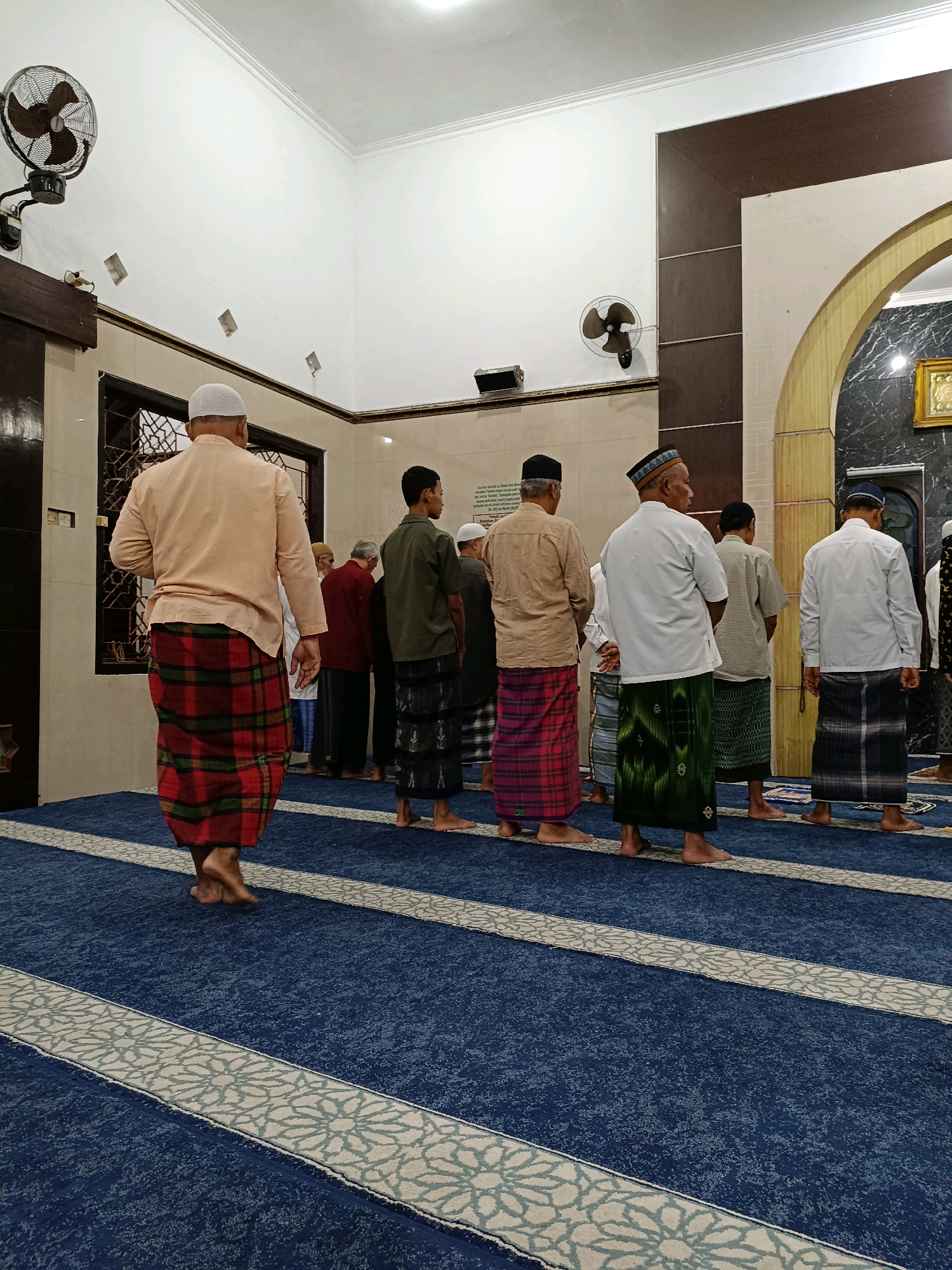 A group of men are standing in a line on a blue carpet with white patterned borders, facing away from the camera. They are in a room with cream-colored walls and a brown ceiling. Some of the men are wearing white caps, and all are wearing sarongs. There is a fan mounted on the wall to the left of the image. The room appears to be a mosque, possibly in Maguwoharjo, Indonesia, given the cultural attire and setting. The lighting is artificial and bright.