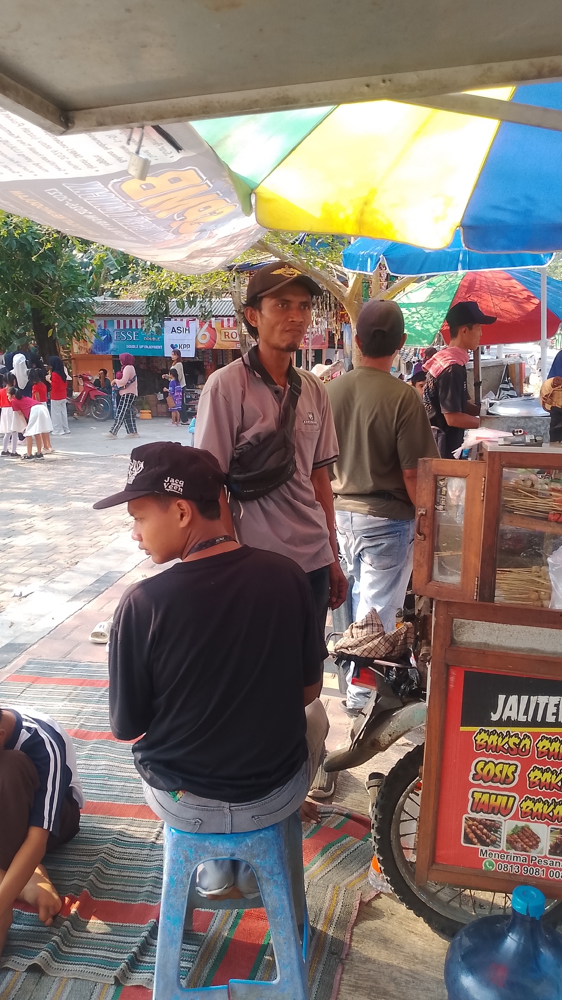 In Karanglangu, Indonesia, a street food vendor operates under a colorful canopy of red, yellow, and blue umbrellas. The vendor displays a variety of grilled food items, including skewers of meat, sausage, and tofu, in a wooden display case mounted on a motorcycle. A sign advertises "JALITEL," followed by descriptions of "Bakso Bakar," "Sosis Bakar," and "Tahu Bakar," and provides a phone number for orders. Several individuals are present, including the vendor, a man in a grey shirt and a black fanny pack, and a young person in a black t-shirt and a baseball cap sitting on a blue stool. In the background, a group of children in uniform, possibly students, are gathered near a red motorcycle. The scene appears to be a bustling outdoor market or gathering place.
