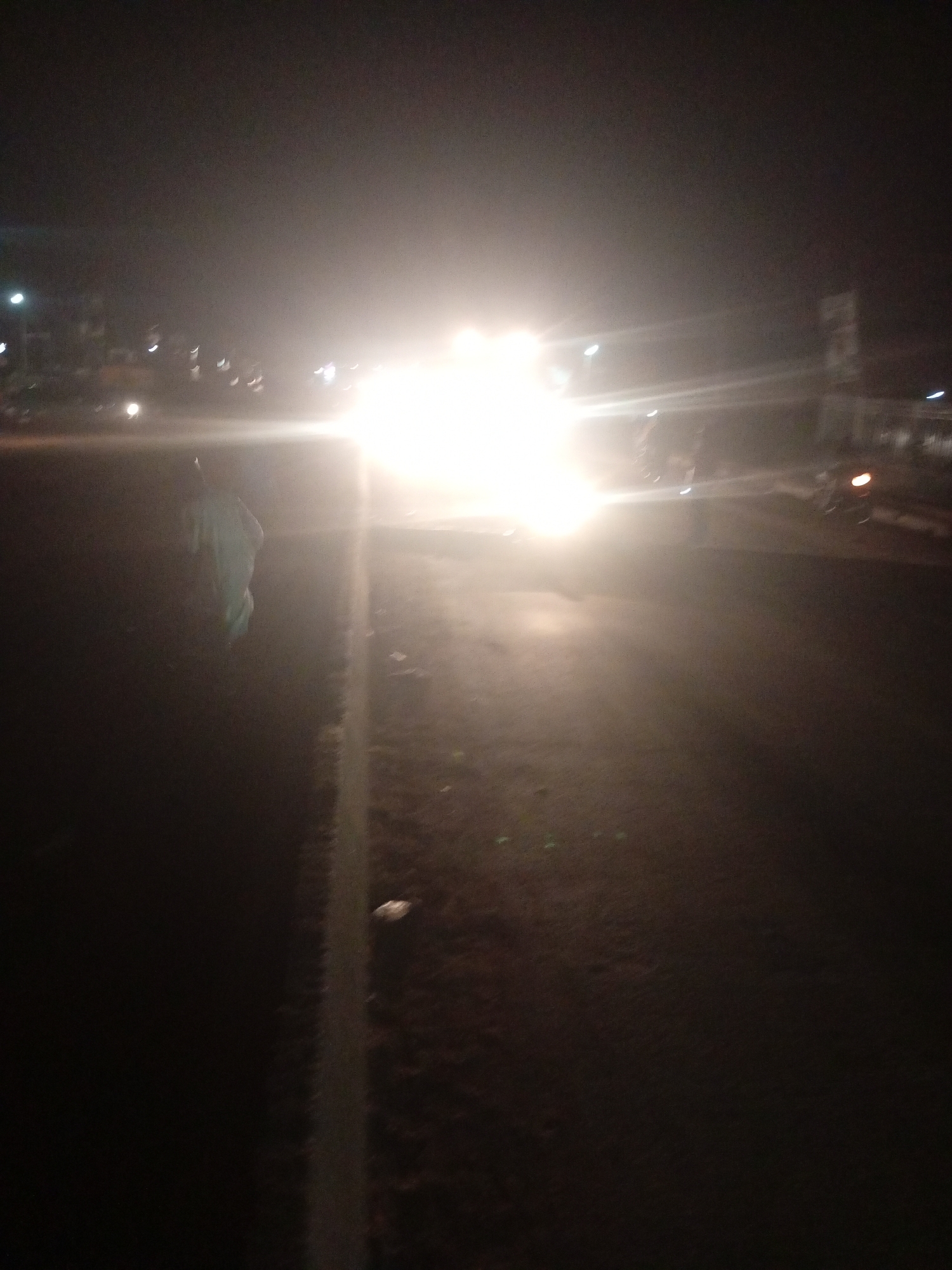 The image depicts a nighttime scene on a paved road in Tundun Wada West, Nigeria, likely a residential or commercial area given the presence of buildings and streetlights. A person, possibly a pedestrian, is visible on the left side of the road, wearing a light-colored garment. The scene is illuminated by a bright, possibly vehicle-mounted, light source, causing significant glare and obscuring details of the surroundings. There are also indications of other vehicles or possibly motorcycles on the road. The road surface appears to be asphalt. The overall impression is of a busy or active road at night, with potential challenges for visibility due to the bright lights and darkness.
