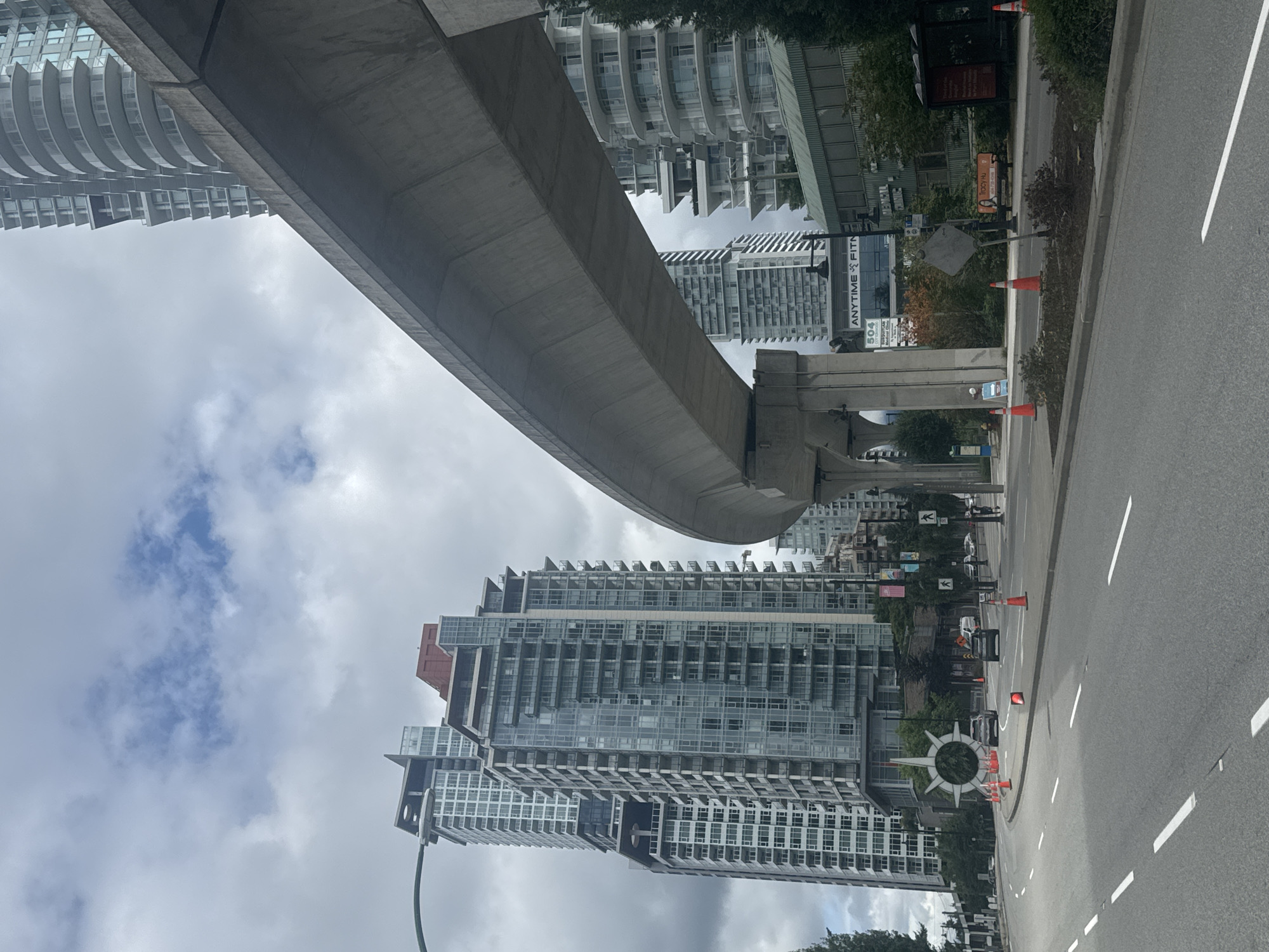 The image depicts an urban street scene in Coquitlam, Canada. In the foreground, a multi-lane paved road with white lane markings stretches into the distance. Along the right side of the road is a sidewalk with trees and traffic cones. A bus shelter is visible on the right side. In the background, a concrete elevated transit line curves across the frame, supported by massive concrete pillars.
Dominating the scene are several modern, high-rise residential buildings with glass facades and balconies. To the left, a particularly tall building features a distinctive red element near its peak. Across the street from the residential towers, a commercial building with a green roof is visible, identified by signage that reads "ANYTIME FITNESS" and "MEDICAL CLINIC".
The sky is overcast with scattered clouds. Several cars are present on the road, including two in the middle distance. Traffic cones are strategically placed along the road and sidewalk, suggesting possible construction or lane closures. In the middle left of the image, a large, circular, spoked sculpture is positioned near the road. People are visible on the sidewalks and in the distance, walking and engaged in everyday activities. The overall impression is a bustling urban environment under a cloudy sky.