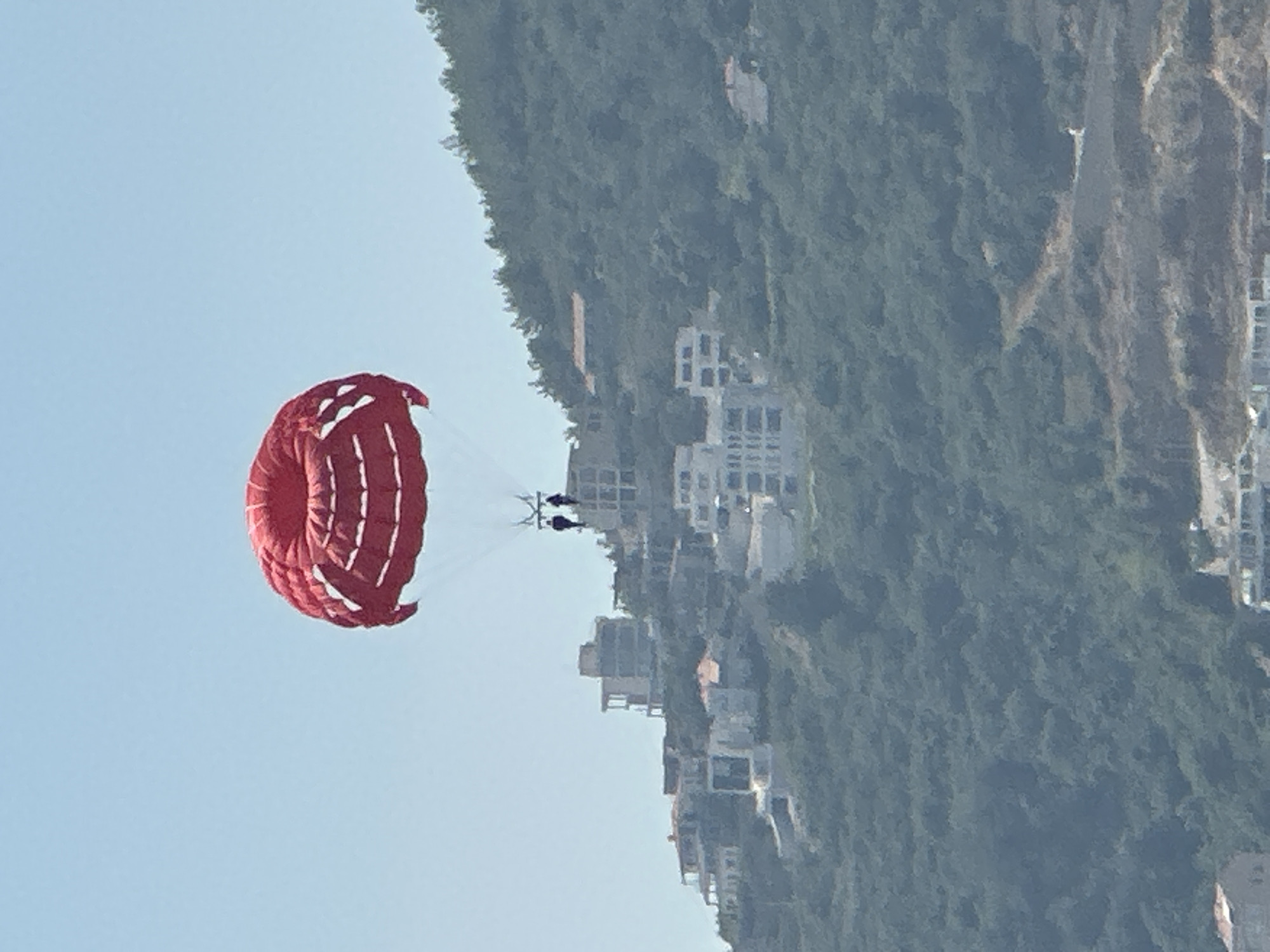 An outdoor, medium shot captures a bright red parasail in mid-air against a clear blue sky. Two people are visible suspended beneath the parasail, attached by ropes. The parasail is positioned in the upper-middle portion of the frame. Below the parasail and extending across the middle ground and background of the image is a densely forested hillside dotted with numerous white buildings of varying sizes and architectural styles. The buildings appear to be concentrated on the upper slopes of the hill, suggesting a coastal town or resort. The vegetation on the hill is predominantly a dark green, creating a textured backdrop for the structures. The overall scene depicts a recreational activity taking place with a backdrop of a hillside urban development.