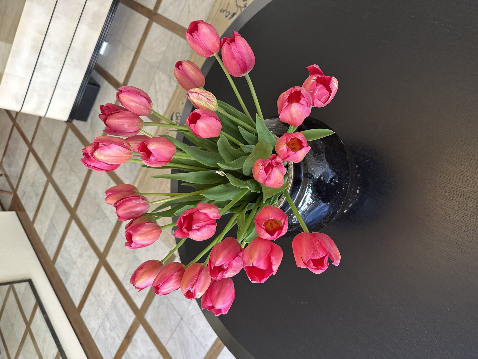 A high-angle shot shows a bouquet of pink tulips arranged in a dark-colored vase with a speckled texture. The vase is placed on a dark, circular table. Part of the surrounding environment includes a geometric patterned floor. The location is not discernible from the image alone.
