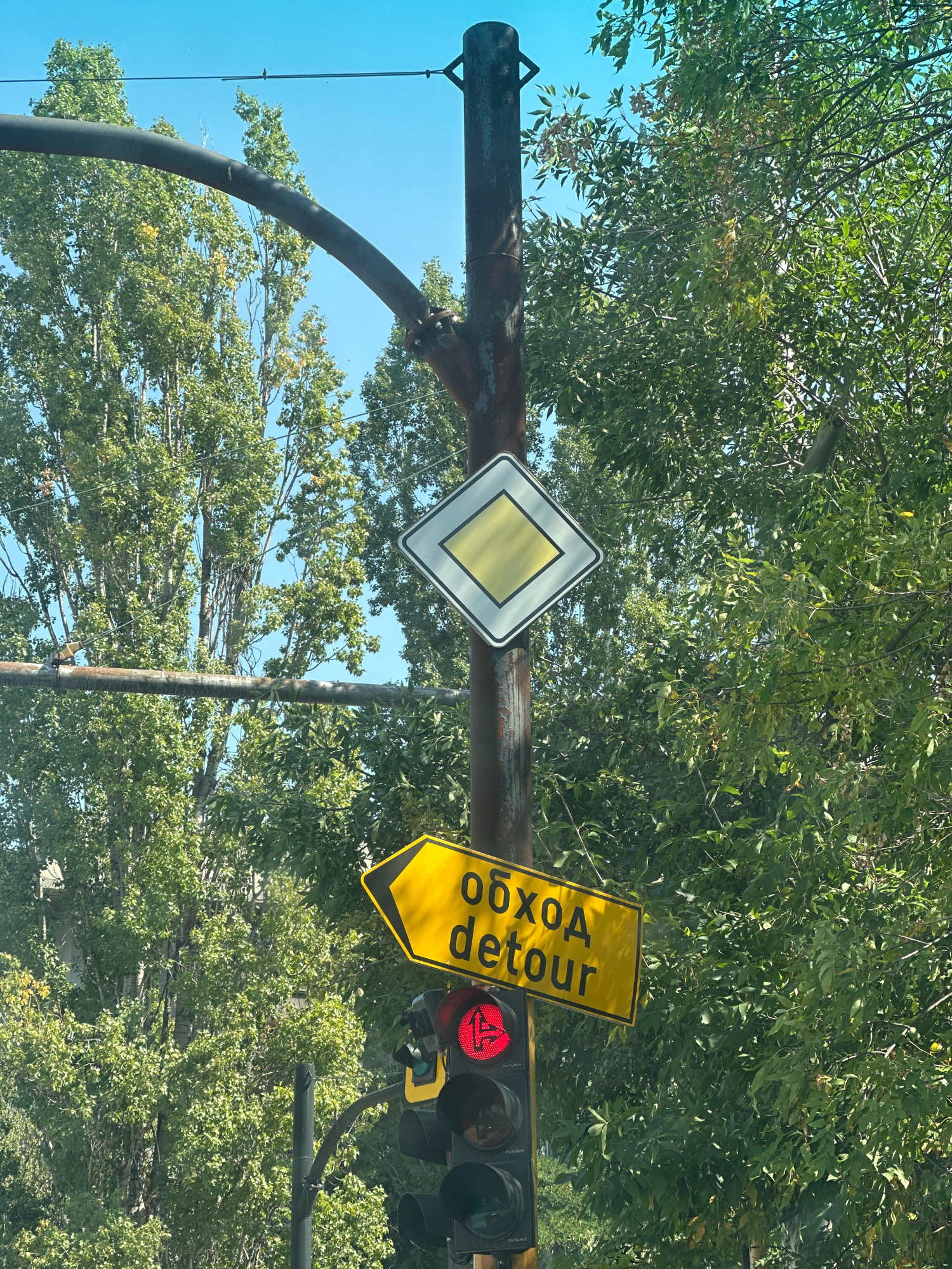 The image depicts a traffic intersection with a traffic light, a main road sign, and a detour sign, set against a backdrop of trees and a clear blue sky. The traffic light shows a red signal. The main road sign is a white diamond with a yellow rhombus inside. The detour sign is yellow with black text in Russian and English, indicating "обход detour" and pointing to the left. The intersection is located in the city of Sofia, Bulgaria. The image is taken from a low angle, suggesting it was captured from a vehicle. The environment appears to be an urban setting with lush green foliage from trees bordering the intersection.