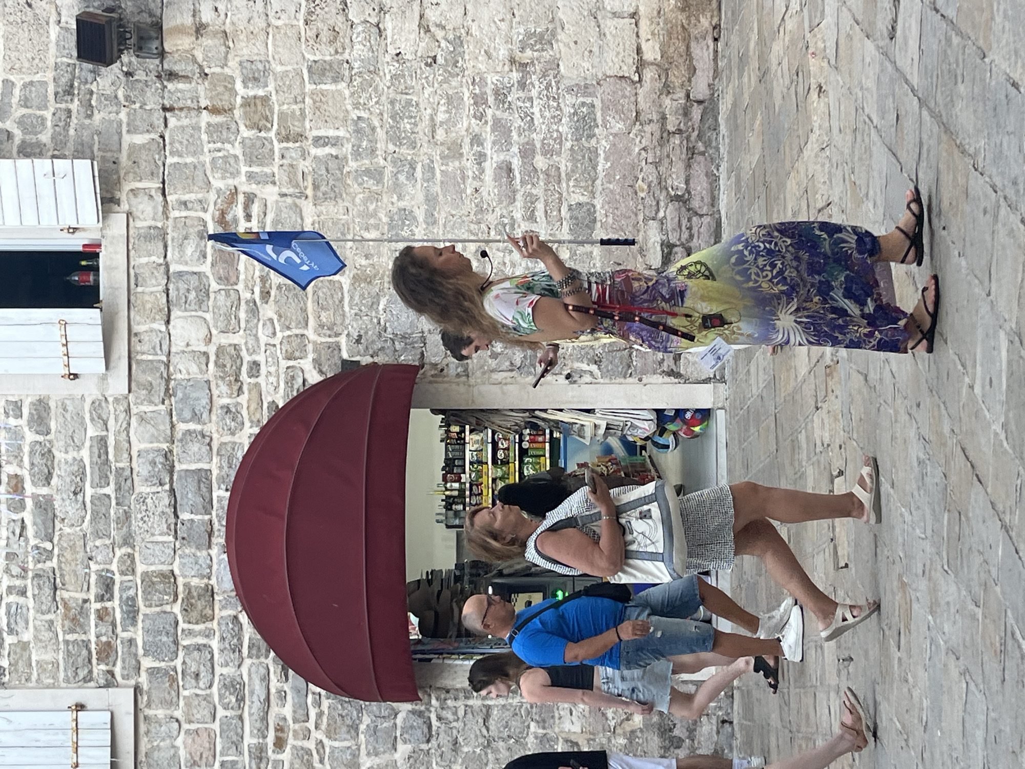 The image depicts a street scene in Budva, Montenegro, likely within the old town given the stone architecture. In the foreground, several people are walking. A woman in a colorful, flowing dress is walking towards the right, holding a mobile phone and wearing a headset. To her left, a woman in a patterned dress carries a white tote bag and also holds a mobile phone. Behind her, a man in a blue t-shirt and denim shorts walks, wearing a black crossbody bag. A woman in a black tank top and denim skirt is to his left. The background features a stone building with a red awning covering an entrance that appears to be a small shop or convenience store. A blue flag with white lettering is visible on a pole near the top of the building. The ground is paved with cobblestones.