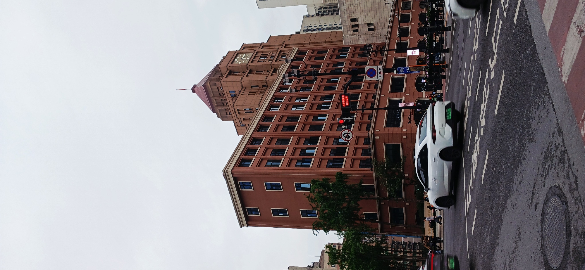 A street-level view in Yuzhong District, China, shows a large, brown brick building with a clock tower rising against an overcast sky. The building features numerous windows. A street runs alongside the building, with a white car in motion, appearing to be traveling past the building. Pedestrians can be seen on the sidewalk. Street signs and traffic lights are present, with one traffic light showing a red signal. Trees line the street. The road surface is wet.