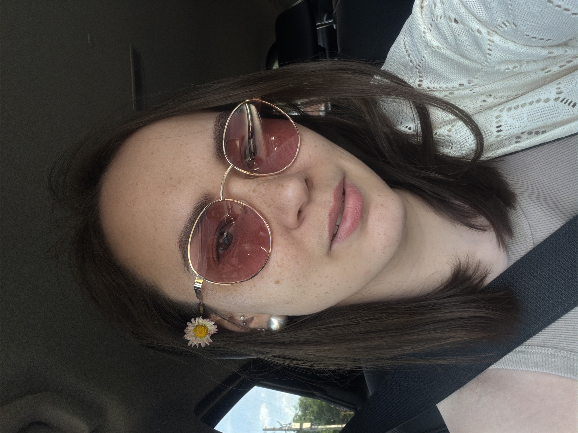 A young woman is in a car, wearing pink-tinted sunglasses and a white daisy tucked behind her ear. She has light skin with freckles, and her hair is dark brown. She is wearing a light-colored top with a white, open-knit sweater over it. A black seatbelt crosses her body. Outside the car window, there is a view of a city street, likely in the daytime, with utility poles and trees visible. Given the prompt, the location is presumed to be Chaikovsky, Russia.