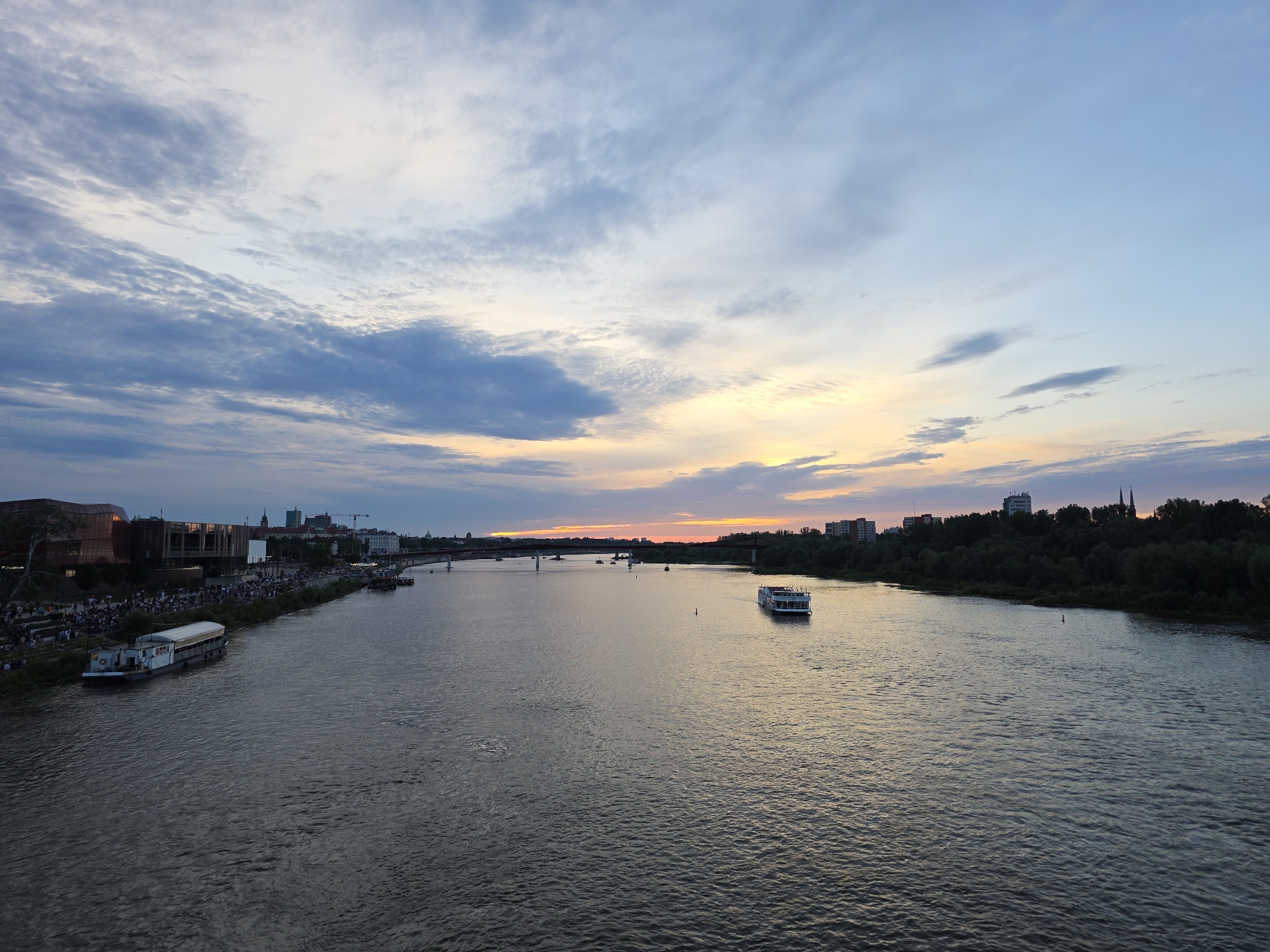 The image captures a wide, scenic view of the Vistula River in Warsaw, Poland, during sunset. The sky displays a gradient of colors, with soft blues and grays at the top, transitioning to a warm orange and yellow hue near the horizon. Wispy clouds are scattered across the sky.
On the left bank of the river, a modern building complex with large windows and a reddish-brown facade is visible. In front of this building, a crowd of people is gathered along the riverbank, with some sitting on terraced steps. A long, white boat is docked along the embankment. Further along the left bank, several other boats are moored, and a bridge spans the river in the distance.
On the right bank of the river, a dense line of trees forms a natural boundary, with buildings and structures of varying heights visible behind the foliage. The river itself is calm, with gentle ripples on its surface reflecting the colors of the sky. Two boats are navigating the water: a larger, white passenger boat with multiple decks is in the middle ground, and a smaller vessel is closer to the right bank. The overall atmosphere is serene and picturesque, indicative of an evening cityscape by the river.