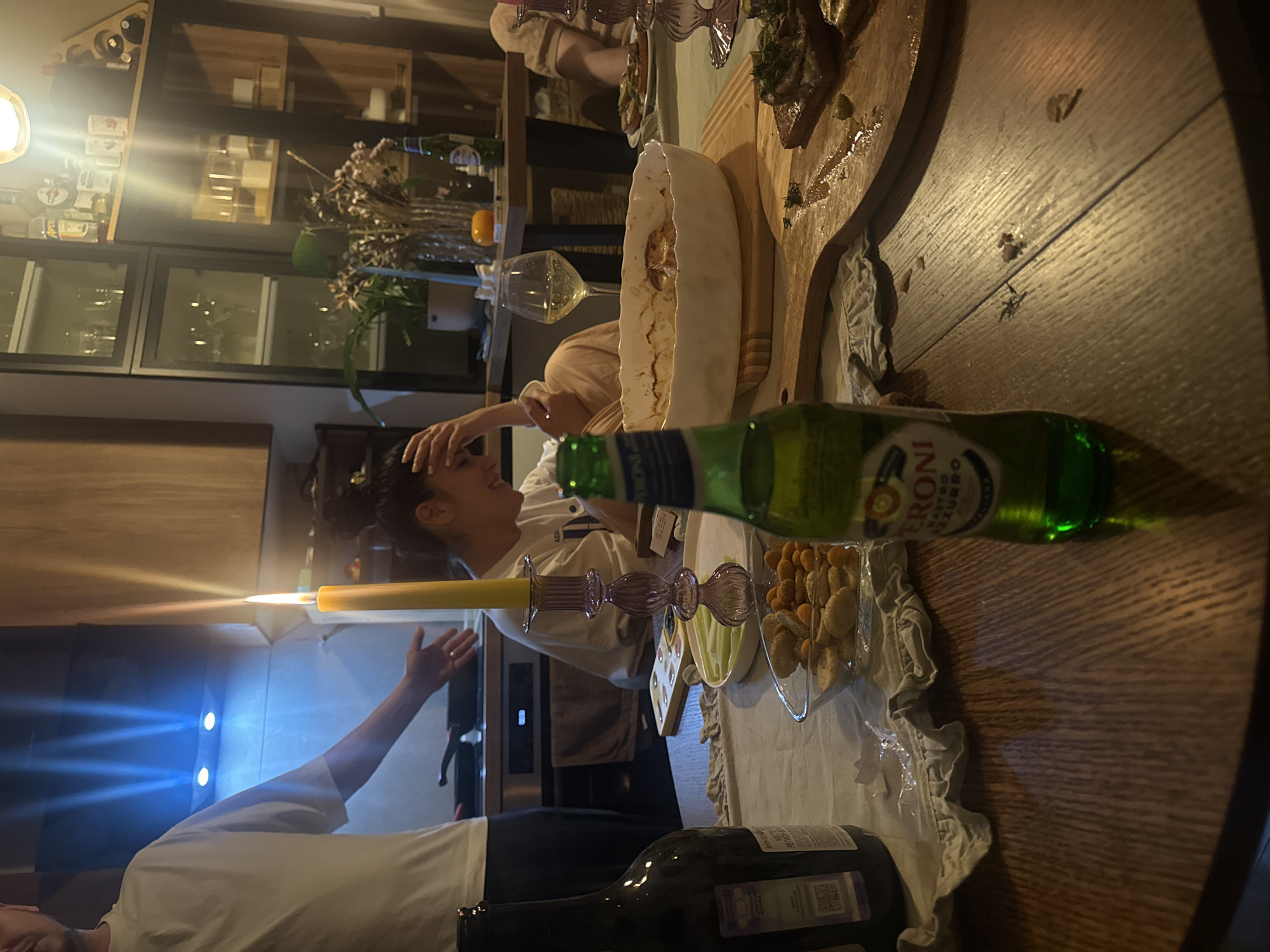 The image depicts an indoor dining scene. Two individuals are visible: one gesturing with an outstretched arm and a second, laughing, with a hand on their head. A table dominates the foreground. On it are an assortment of items including a lit candle in a glass holder, an unopened bottle of Peroni beer, a wooden board with food, a bowl, glassware, a dish with snacks, and a bottle of wine. The background features a kitchen area with cabinets, appliances, and decorative elements such as floral arrangements.
The information provided suggests the location is Razvilka, Russia, but the image itself offers no means to confirm this.
