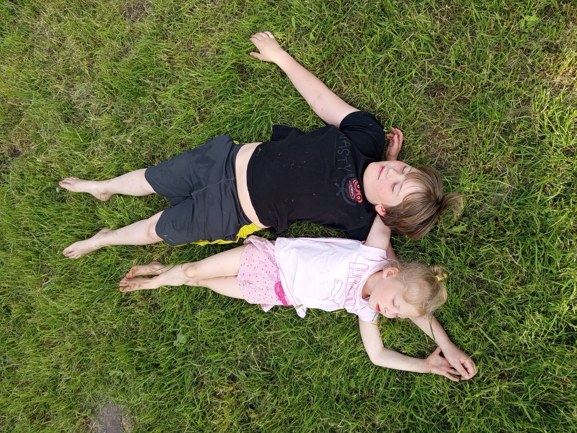 This overhead image, captured in Ostrovtsy, Russia, features two children, a boy and a girl, lying side-by-side on a vibrant green lawn. Both children have their eyes closed, conveying a sense of peace and relaxation, possibly sleeping or resting.
The boy, positioned slightly above and to the left, wears a black t-shirt with the word "NASTY" printed on it, and dark shorts with a visible yellow trim along the waistband. He has brown hair and is barefoot, with some soil on his feet and lower legs. His left arm is extended upwards, while his right arm rests bent by his head.
The younger girl lies below and to the right of the boy. She wears a light pink t-shirt featuring the "PUMA" logo and text, paired with pink shorts adorned with white polka dots. Her blonde hair is tied back with a yellow hair tie, and like the boy, she is barefoot, with dirt visible on her feet and legs. Her arms are bent, resting on the grass near her head, and her right hand appears to be lightly grasping some blades of grass.
The scene suggests a warm, sunny daytime setting, evidenced by the bright natural light and the children's light attire. The lush, green grass fills the frame, indicating an outdoor, natural environment.