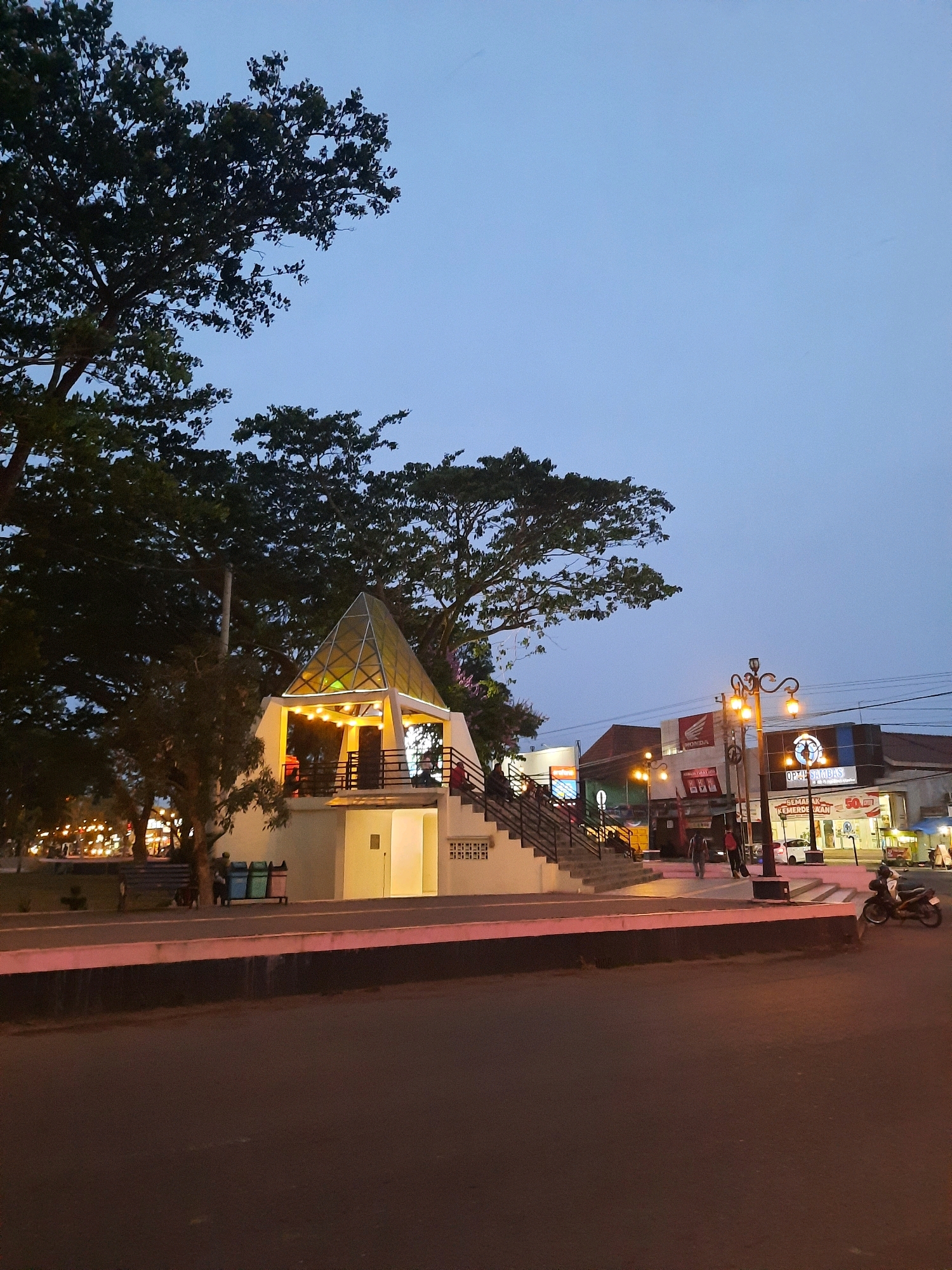 The image depicts a dusk scene in the city of Kutoarjo, Indonesia. In the foreground, a dark asphalt road occupies the lower portion of the frame, bordered by a raised concrete barrier with a pinkish-white stripe. To the left of the road, a paved walkway runs parallel, with a dark-colored bench and three trash bins. Beyond the walkway is a raised platform with a white structure topped by a glass-paneled, pyramid-shaped roof illuminated with yellow string lights. A set of stairs leads up to this structure. Several trees with dark foliage are visible, with one large tree dominating the left side of the image and another on the right.
In the mid-ground, across the platform, a street scene unfolds. Buildings with signage are visible, including one with a "HONDA" logo and another with "OPTIC SAMBAS". Streetlights with ornate metal poles cast a warm glow. Several people are walking on the sidewalk, and a motorcycle is parked on the right side of the frame, with its rider seated. The sky is a clear, pale blue, indicating the transition from day to night. The overall impression is one of a public space in a town or city at twilight, with a mix of natural elements and urban infrastructure.