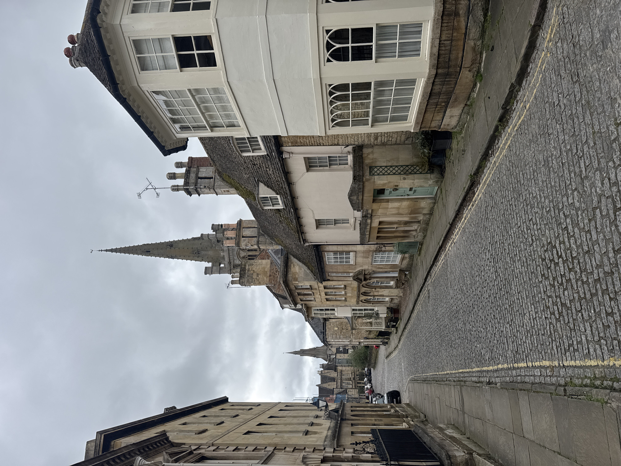 The image captures a narrow, cobblestone street lined with buildings of varying heights and architectural styles. A church steeple rises prominently in the background against a cloudy sky. The buildings are made of stone and brick, with windows and doors. There is a yellow line painted on the edge of the cobblestone street. People and vehicles are visible in the distance, suggesting a lively urban environment. The location is South Kesteven, United Kingdom.