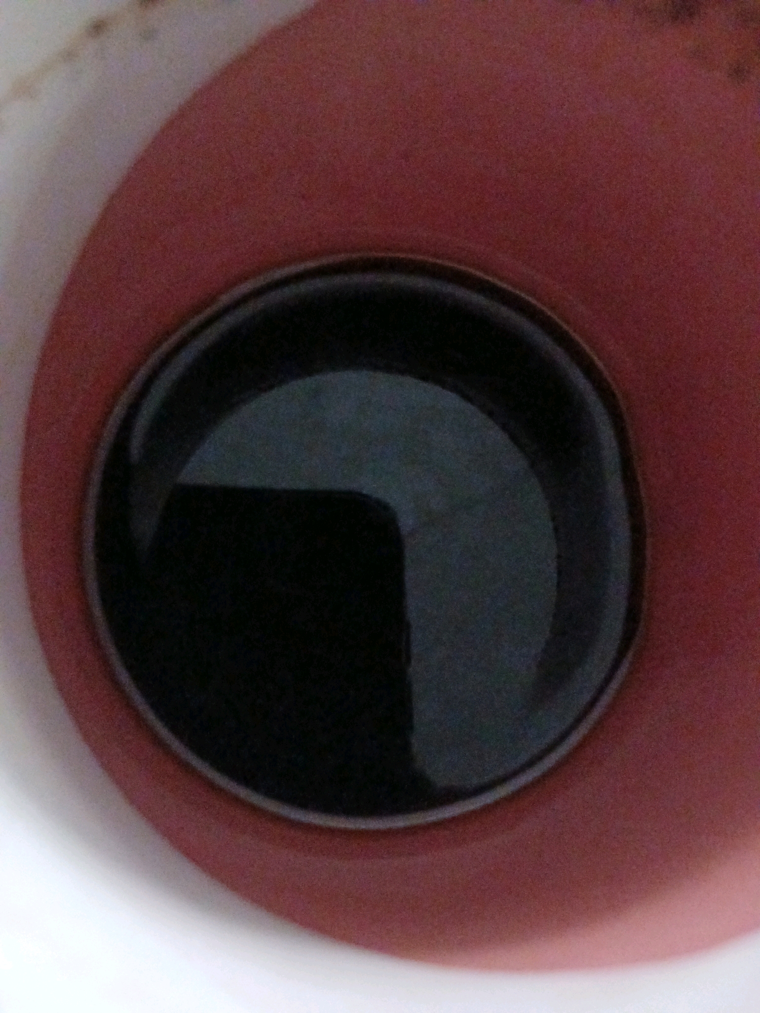 The image shows the surface of a dark liquid, likely coffee, contained within a red mug. The mug appears to be made of ceramic and has a slightly textured surface. Reflections are visible on the surface of the liquid, suggesting the presence of overhead lighting. The background is blurred and predominantly red, consistent with the interior of the mug. No specific location in Kademangan Kulon, Indonesia, can be identified from this image, as it focuses solely on the contents of a mug. There are no people, actions, or interactions depicted.
