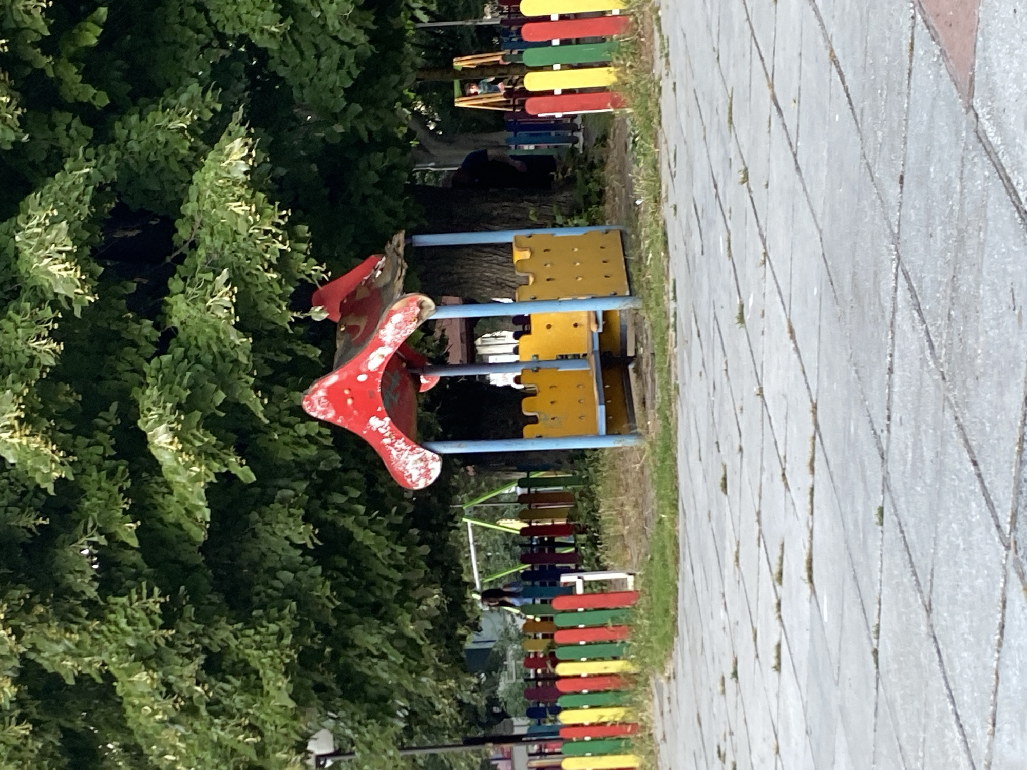 The image depicts a small, dated playground area situated in the city of Varna, Bulgaria. In the foreground, a paved area made of light gray square tiles, some of which have sparse grass growing between them, leads towards the playground. A colorful, low fence with alternating red, yellow, green, and blue vertical posts encloses a section of the playground. Within this fenced area, a red and yellow playhouse-like structure stands, supported by blue poles. The playhouse roof is red with white circular accents, and its side panels are yellow with cut-out shapes resembling castle battlements. A bench is visible within the structure. Behind the playhouse, a large tree with abundant green foliage provides shade. In the mid-ground, beyond the colorful fence, a swing set with green swings can be seen. Two individuals are partially visible; one woman in a light blue top stands behind the colorful fence, near the swing set. Another person, wearing a dark shirt, is obscured by the trunk of the large tree, near the playhouse. The overall scene suggests a public park or a communal play area, likely in a residential district of Varna.
