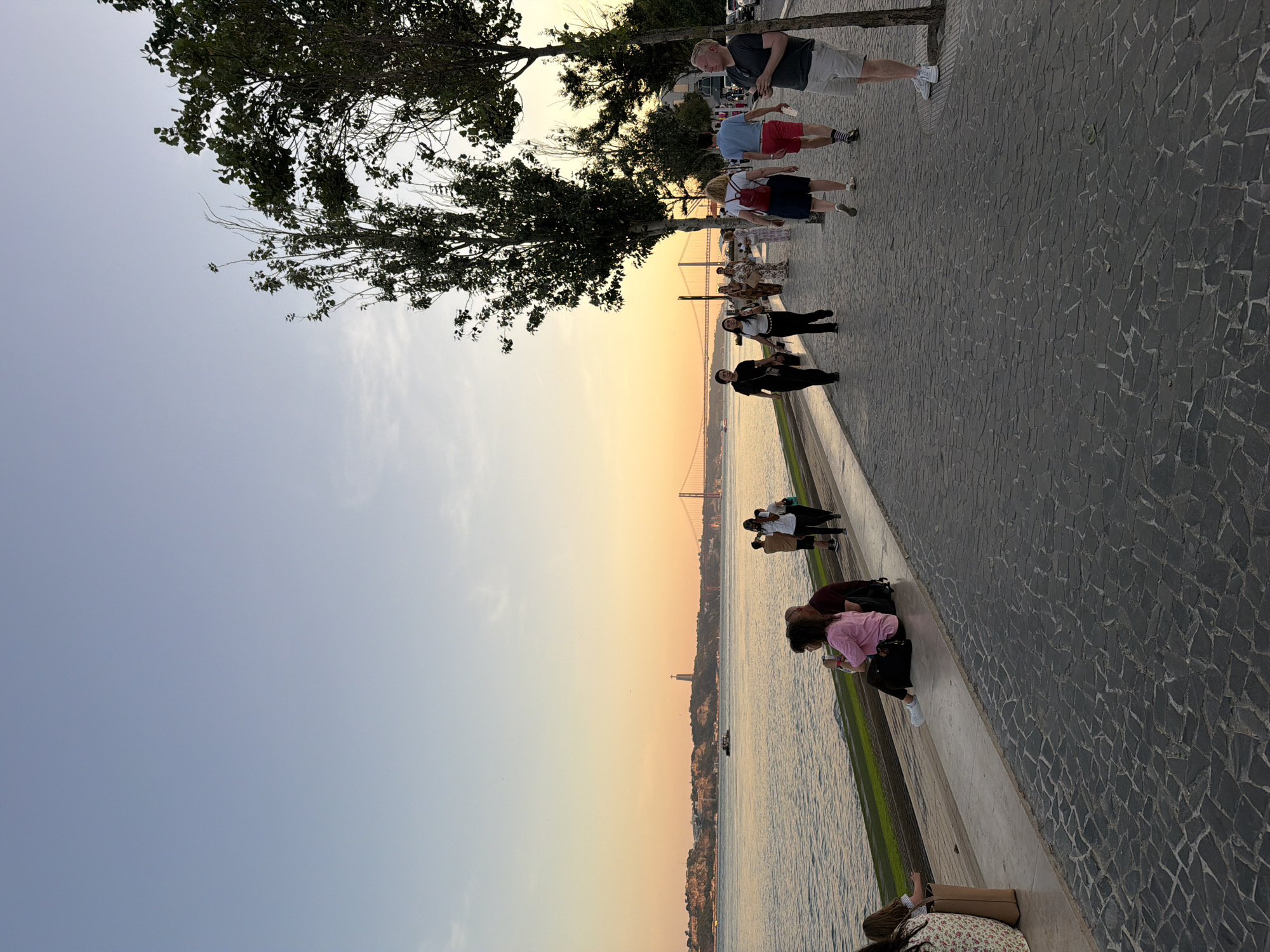 The image depicts a waterfront promenade in Lisbon, Portugal, during sunset. The sky transitions from a pale blue to a warm orange hue near the horizon. A wide river, likely the Tagus River, occupies the left side of the frame, with a boat in the distance. Across the river, the city skyline is visible, including a prominent monument on a hill and a large suspension bridge.
On the right side of the image, a paved pedestrian walkway runs alongside the river. Several people are walking along the path, while others are seated on a low wall bordering the river. The walkway is paved with dark cobblestones. Trees with green foliage line the promenade, casting shadows on the ground.
The people visible are casually dressed and appear to be enjoying the evening. One person in the foreground on the right is holding a phone. Another person, further down the walkway, is wearing a backpack. On the riverbank, two individuals are seated on the wall, facing the water. The overall atmosphere is relaxed and scenic, characteristic of a popular public space in Lisbon.