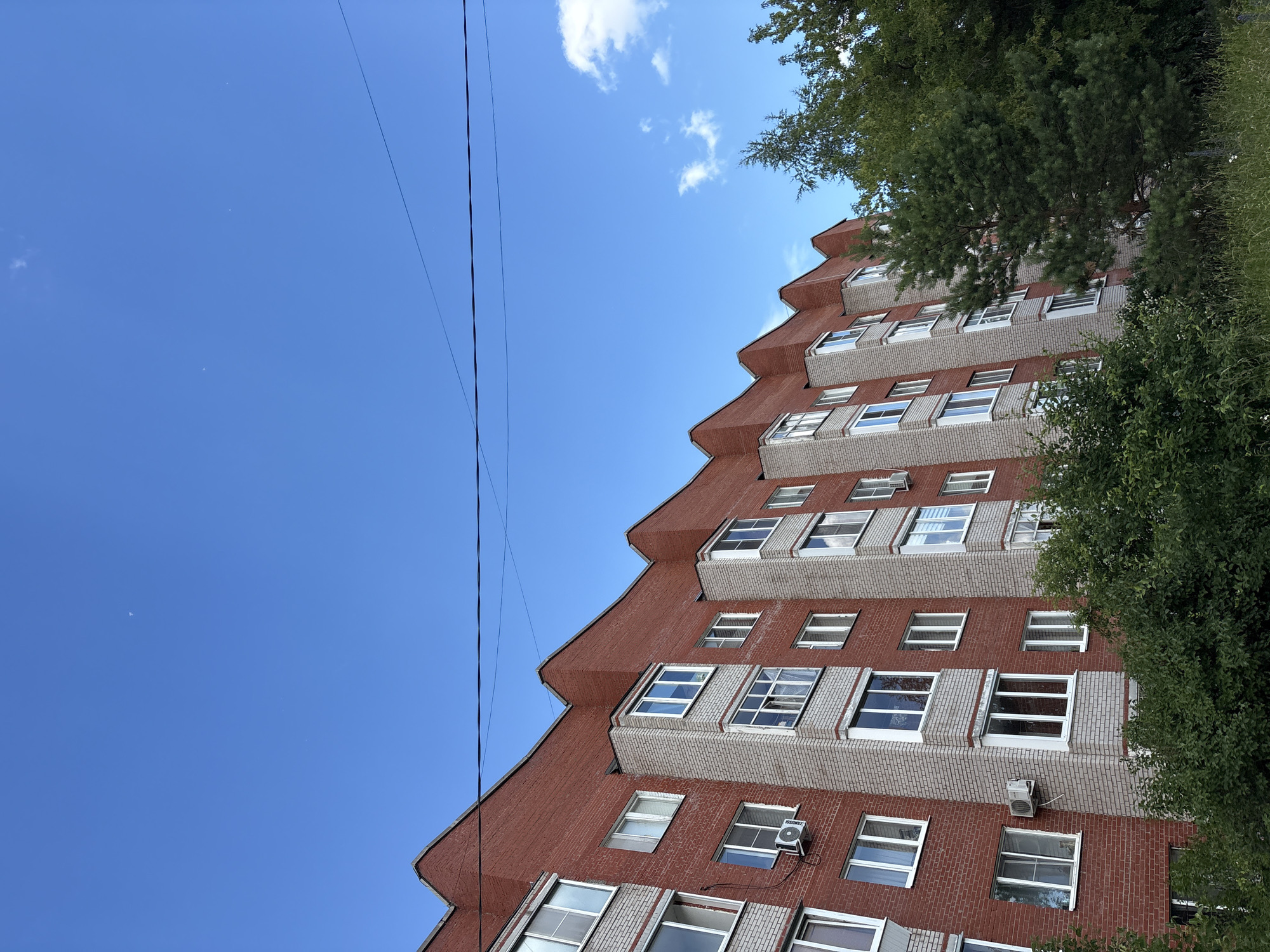 The image is an exterior, low-angle shot of a modern apartment building with a distinctive zigzag facade, set against a clear blue sky. The building is constructed with reddish-brown brick and light grey brickwork, featuring numerous windows, some with open shutters. Several air conditioning units are visible mounted on the exterior walls. Overhead power lines crisscross the sky on the left side of the frame. Lush green trees and foliage frame the right side of the building. The scene suggests a bright, sunny day, possibly in the late morning or early afternoon, given the quality of light and clear sky. There is no visible text in the image. The location is identified as Chaikovsky, Russia. No people or specific activities are depicted, creating a static scene focused on the architecture and its surroundings.