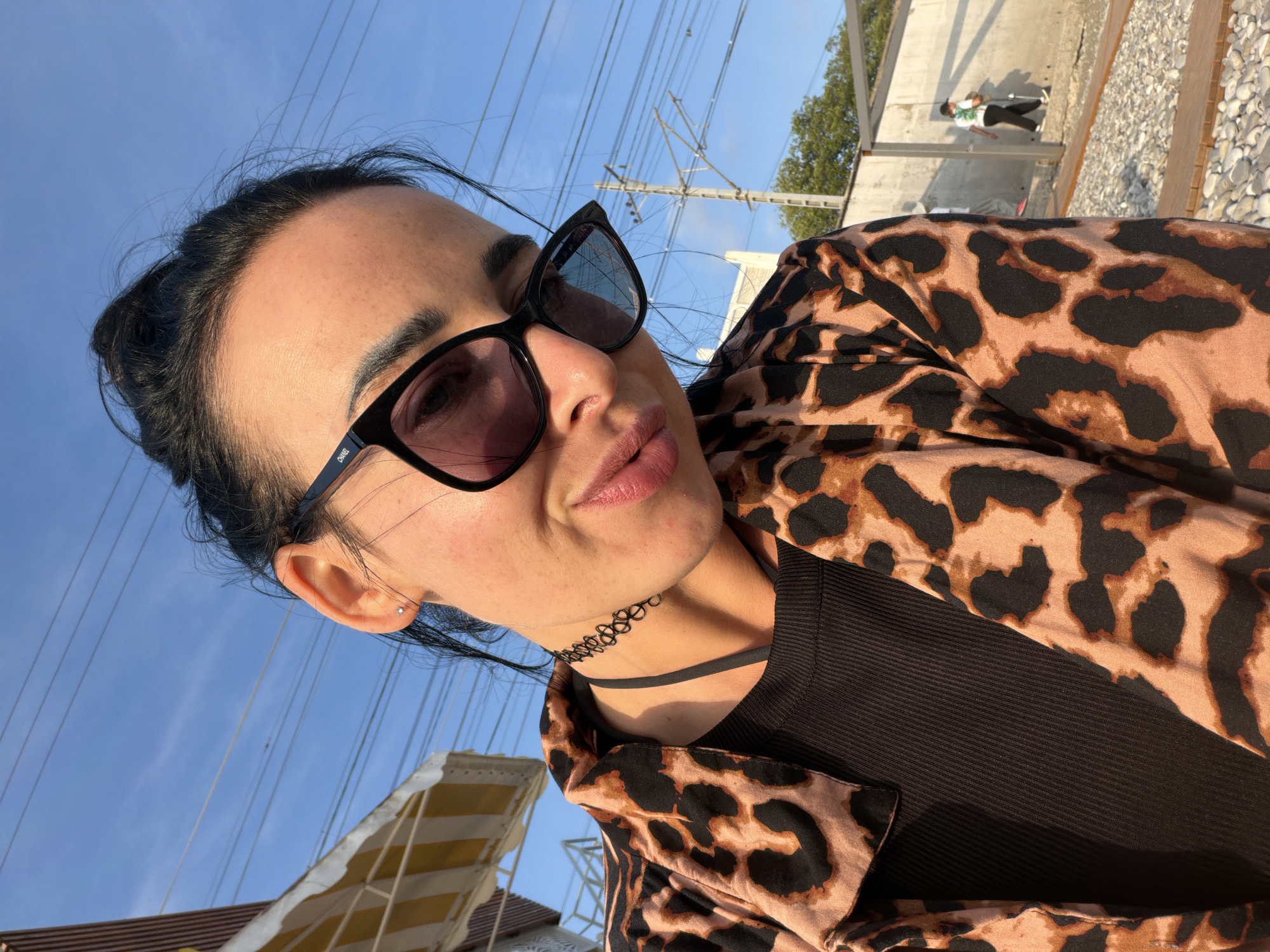 In an outdoor setting under a clear blue sky, a young woman with dark hair pulled back in a bun poses for a selfie. She is wearing large, dark-rimmed sunglasses that obscure her eyes, a black choker necklace, and a black ribbed top. Over the top, she has on a leopard-print jacket. Her head is tilted slightly, and her lips are puckered. In the background, there are power lines strung between poles, a striped awning of a beach umbrella, and a concrete wall. A person in a white shirt and dark pants is walking away from the camera in the distance. The ground is covered with small stones and wooden planks.