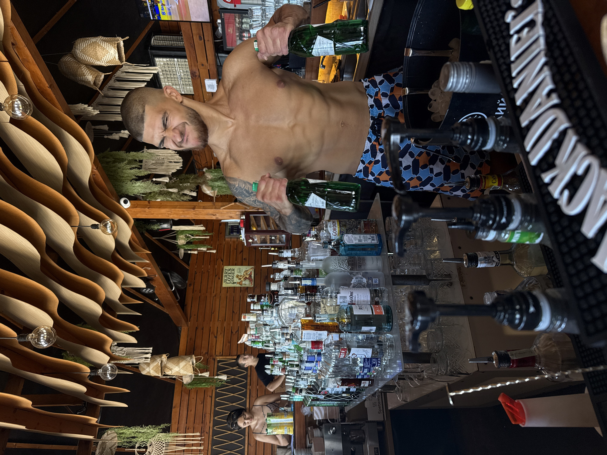 The image shows a man holding two green bottles, appearing to be a bartender at a rustic bar. He is shirtless, wearing patterned shorts, and has a strained expression. The bar is decorated with wooden paneling, hanging baskets, and exposed light bulbs. Behind him, a collection of bottles of alcohol is neatly arranged on shelves and a countertop, including brands like Jack Daniel's, Ketel One, Belvedare, and Gordon's. Several glasses are also visible. In the background, another person, a woman, is partially visible, also seeming to work at the bar. A sign on the wall reads "Jerry Loves Ginger." The setting suggests a lively atmosphere, possibly during a busy service or a break. The lighting is warm and ambient, hinting at an evening or late afternoon time. The overall impression is one of a casual, well-stocked bar environment.