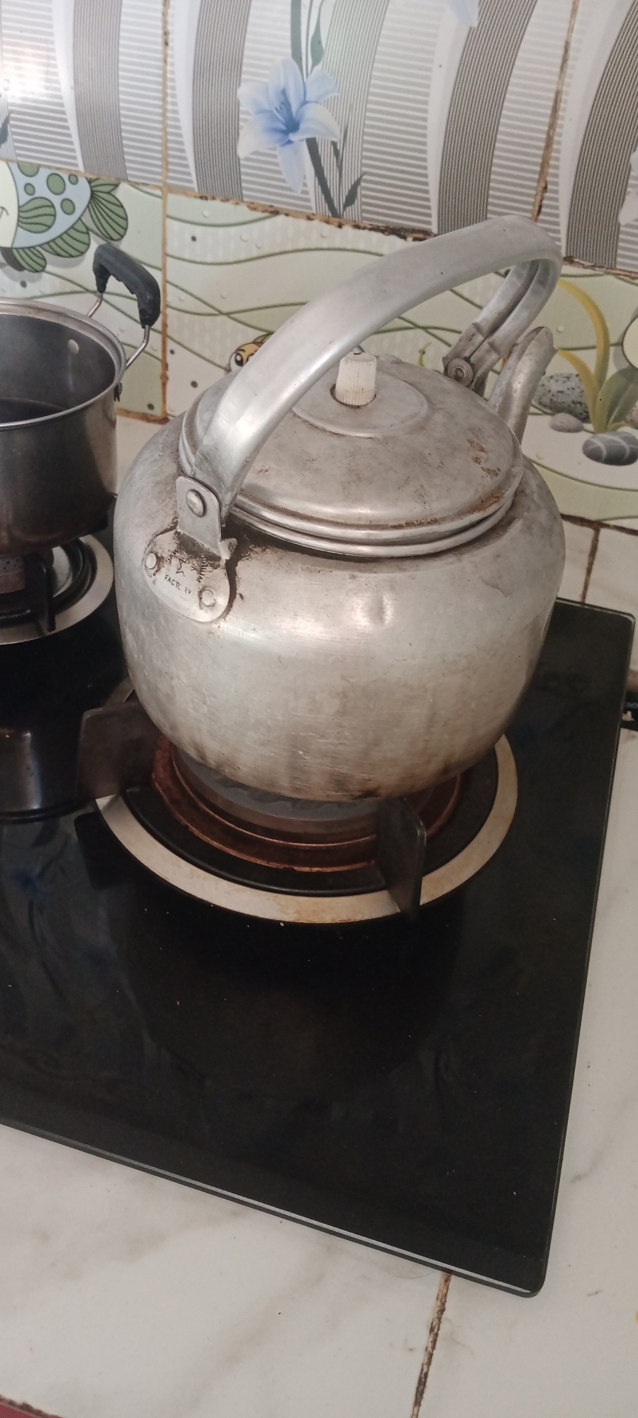 A medium shot captures a silver kettle sitting on a black glass stovetop in a kitchen. The kettle has a prominent handle and a white knob on its lid. To the left of the kettle, a stainless steel pot with a black handle is partially visible. The stovetop is placed on a white countertop. The background features tiled walls with a decorative pattern of grey stripes and a light blue flower. The scene appears to be located in a home kitchen in Rengel, Indonesia.