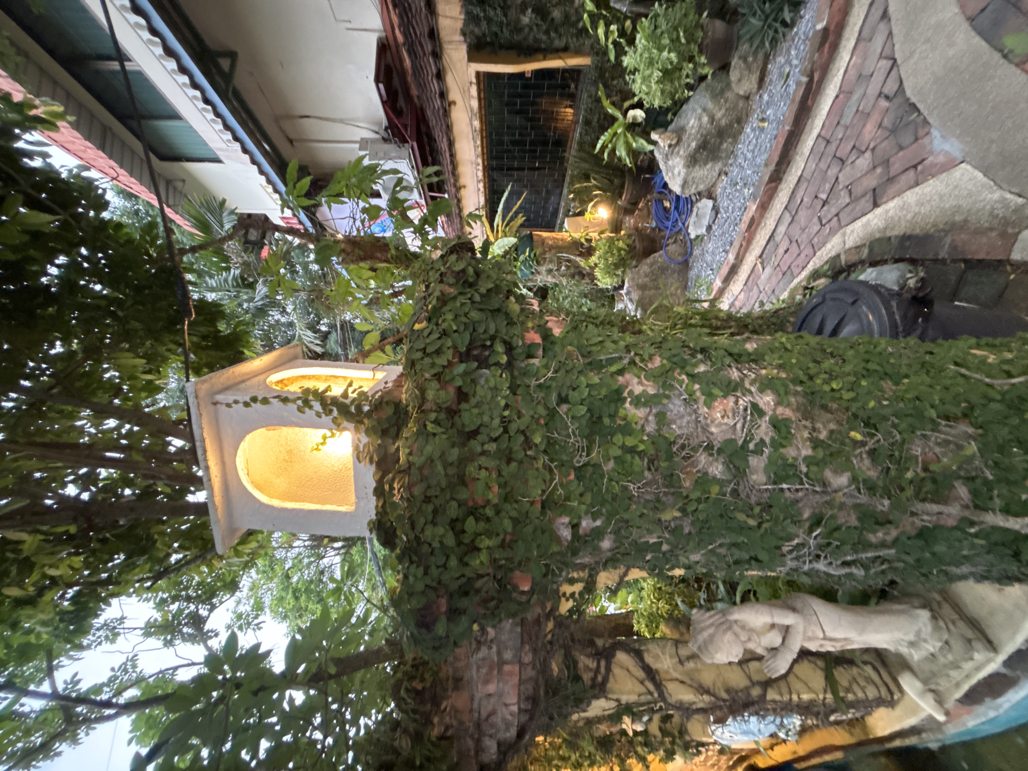 The image depicts a dimly lit outdoor scene in Chalong, Thailand, likely in a private residence or garden. In the foreground, a brick pillar is heavily covered in green ivy, with a small, white, arched structure on top emitting a warm yellow light. To the left, a pale stone statue of a woman in prayer is visible, partially obscured by the ivy. A pathway made of red bricks and grey gravel winds through the scene, leading towards a building in the background. This building has a tiled roof, multiple windows with dark panes, and white walls. Various potted plants and large rocks are scattered along the pathway and around the building. A blue hose and a black trash bin are also present. The overall atmosphere is serene and somewhat mysterious, with the natural elements contrasting with the man-made structures.