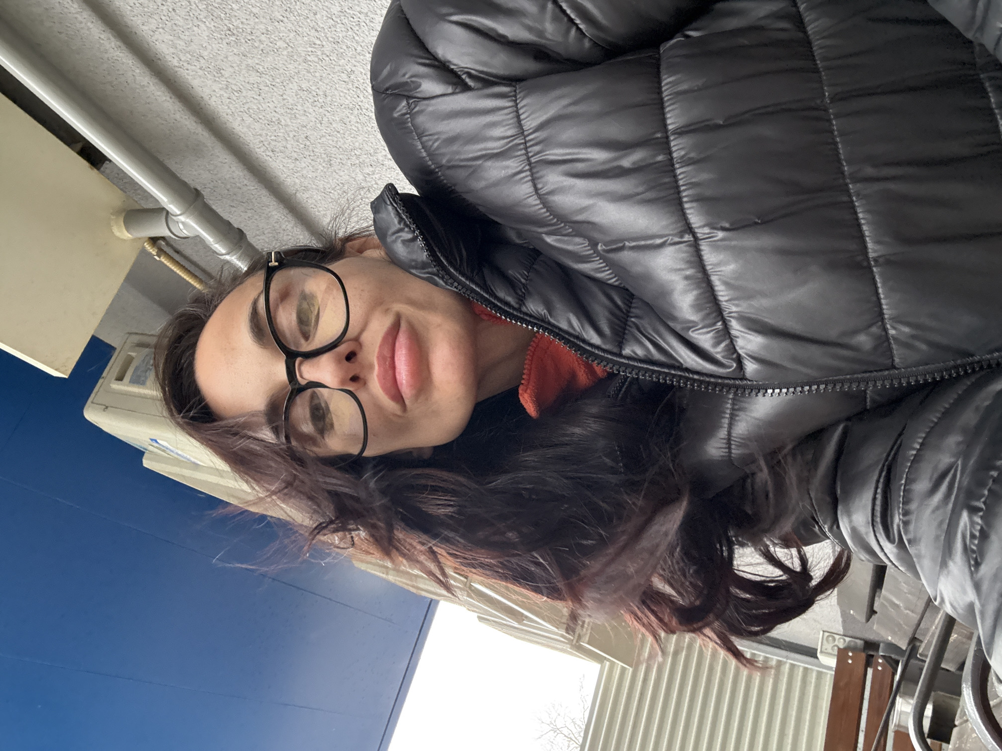 A woman with dark hair and glasses is wearing a black puffer jacket. The background includes a blue wall, a white building, and metal pipes. The location is Pleven, Bulgaria.