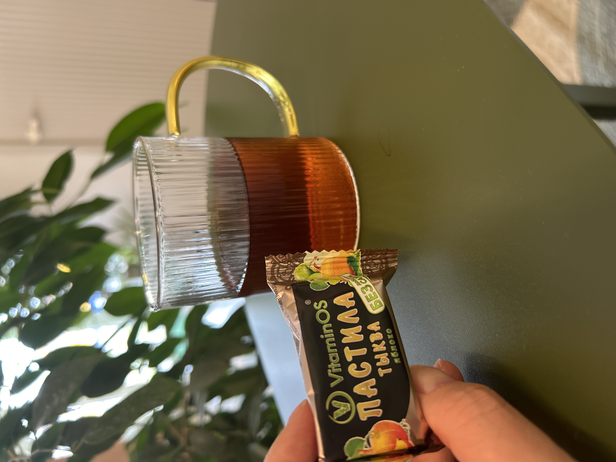 An individual is holding a wrapped candy in their left hand, positioned in the foreground of the image. The candy wrapper is black with colorful illustrations of fruit and text. Behind the candy, a clear glass mug filled with a dark amber liquid, likely tea, is visible. The mug has a ribbed texture and a golden handle. The setting appears to be indoors, with a dark green table surface in the foreground. In the background, blurred green foliage suggests the presence of plants, and a light-colored vertical surface, possibly a wall or door, is also visible. The scene is well-lit, possibly by natural light. No specific location in Medvedevo, Russia, is indicated by any visible elements in the image.