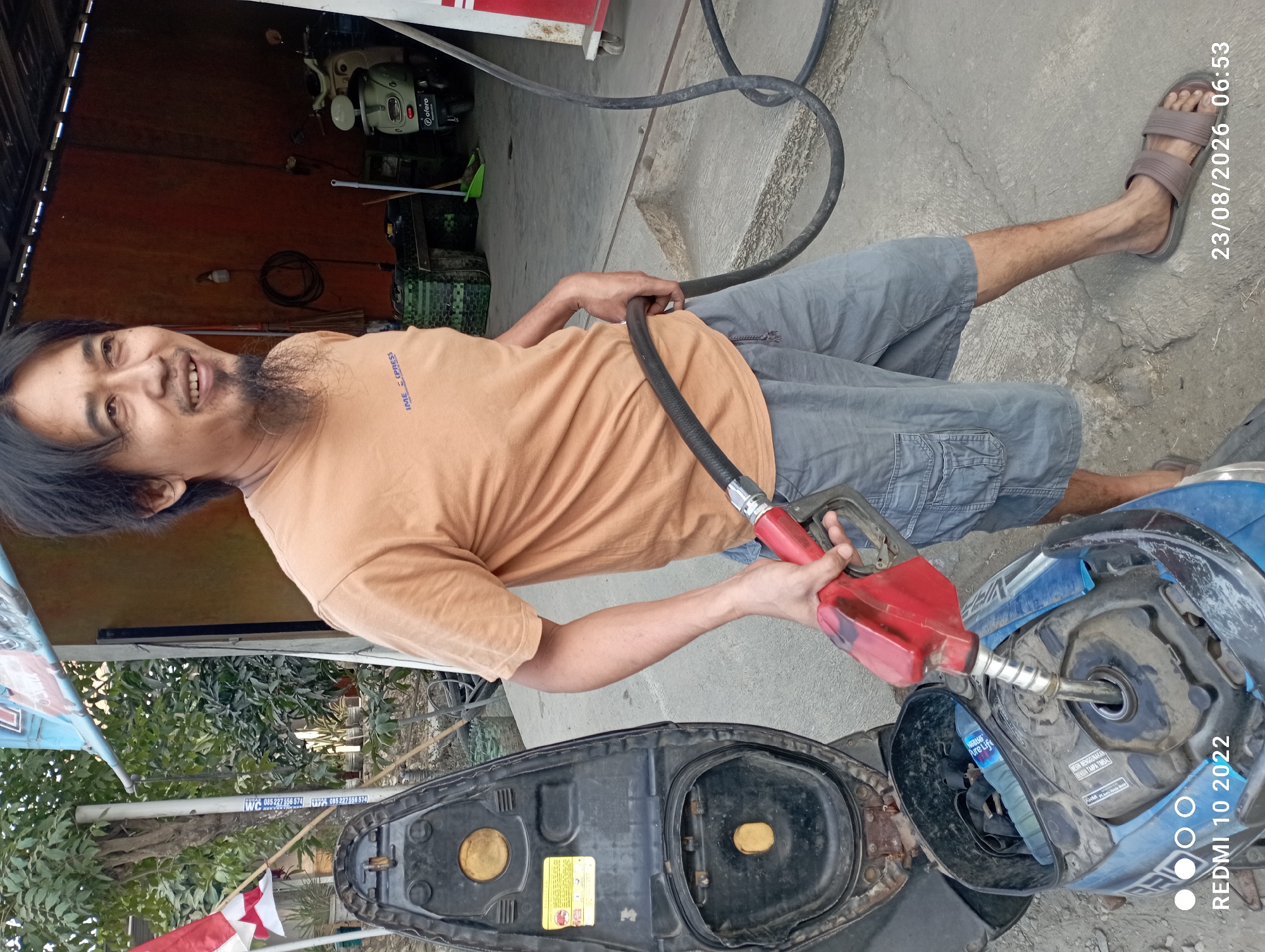 A man with dark hair and a goatee is filling a blue motorcycle with gasoline in Dukuhseti, Indonesia. He is wearing a light brown t-shirt and grey cargo shorts, and brown sandals. He is holding a red gas nozzle connected to a black hose. The motorcycle is partially disassembled, with the seat lifted. In the background, there is a gas pump and a parked scooter. The ground is concrete, and there are trees and foliage visible. A red and white flag is also visible on the left side of the image. The timestamp in the bottom right corner indicates the photo was taken on 23/08/2026 at 06:53.