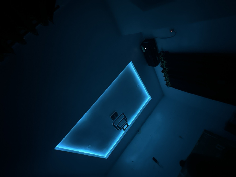 A dim room, illuminated by a rectangular recessed ceiling light that emits a vibrant blue glow, highlights an unusual overhead fixture that looks like a printer. Above and right is a dark projector, partially obscured. To the right, in the same area, an equally dark curtain hangs. Other features, like furniture and additional decor, are indistinguishable due to the poor lighting conditions.
