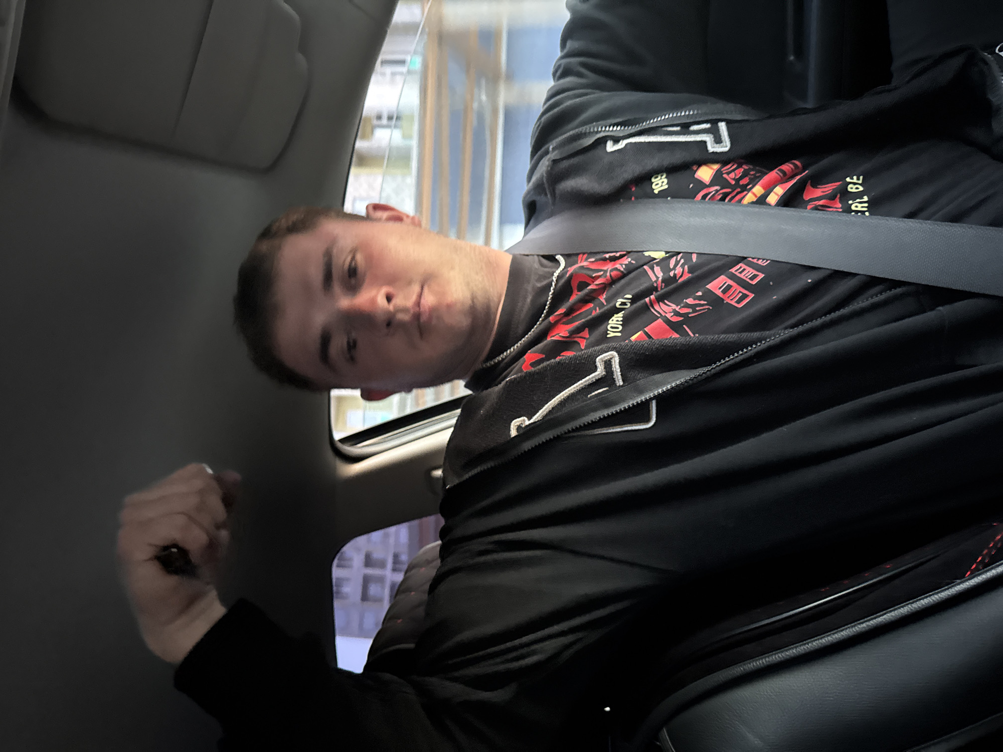 A Caucasian man is seated in a vehicle. He is wearing a black jacket over a black and red patterned shirt with a silver chain. A gray seatbelt crosses his chest. He is looking at the camera with a neutral expression. His left hand is visible, held in a fist near the car door. The background through the window shows blurred city buildings. It is not possible to determine the location as Yakutsk.
