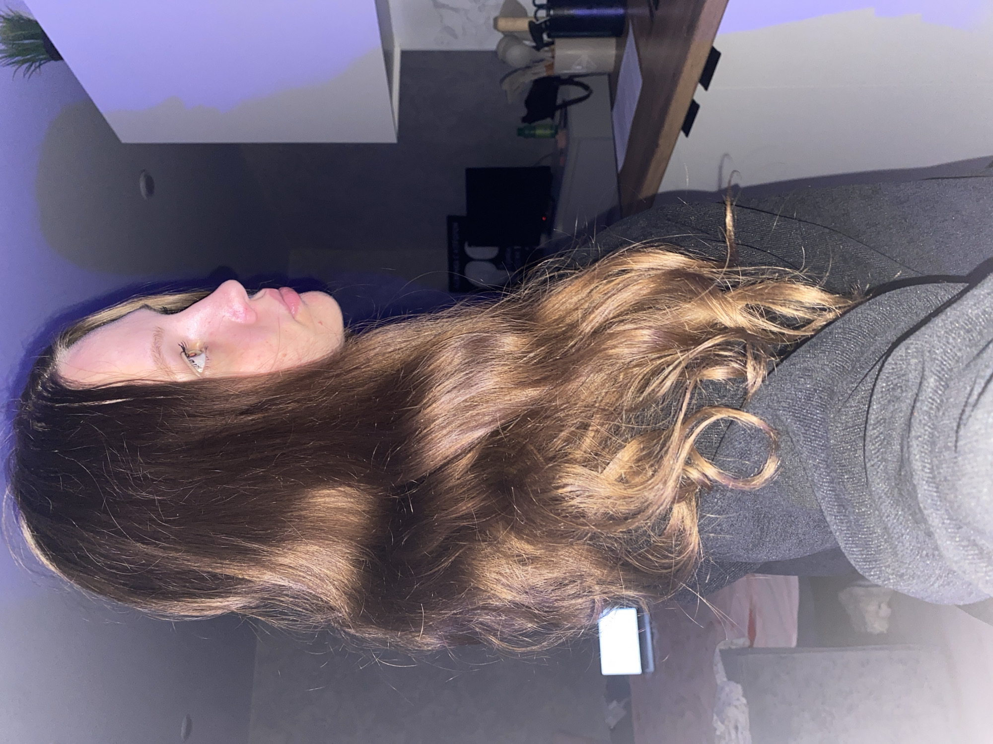 A woman with long, wavy, brown hair is lying down. She is wearing a dark gray jacket. The background is a dimly lit room with a white ceiling and walls. A shelf with various items is visible in the upper portion of the frame. The location is not identifiable as Izhevsk, Russia from the visual information provided.
