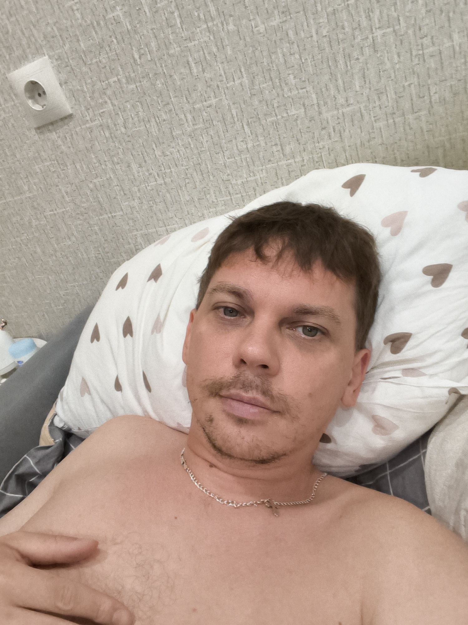 A shirtless man with short brown hair and a mustache lies on a pillow with a heart pattern, looking at the camera. He is wearing a silver chain necklace. The background consists of a textured wall with a white electrical outlet. The scene appears to be indoors, possibly a bedroom. There is no information to suggest this location is in Chelyabinsk, Russia.