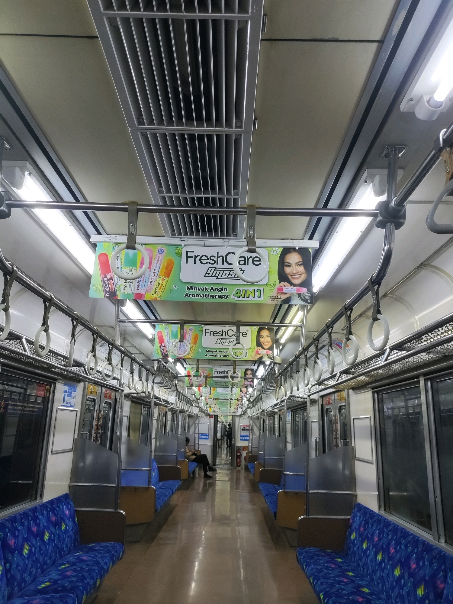 The image captures the interior of a train carriage, looking down the aisle toward the front. The carriage is equipped with rows of blue upholstered seats on both sides, separated by metal dividers. Above the seats, a network of metal handrails and straps is visible, with several white plastic handles hanging down. Advertising posters featuring a woman and product branding, including "FreshCare," are displayed overhead. Fluorescent lights illuminate the interior. The floor is a polished, light brown material. Towards the front of the carriage, a person is seated on the left side, facing away from the camera. The overall environment suggests a public transportation setting, likely within a city. There are no specific visual cues within the image to confirm the location as Telagaasih, Indonesia.