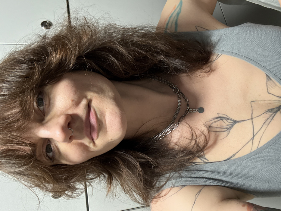 The image displays a close-up, chest-up portrait of a female subject looking directly at the camera with a slight smile. She has shoulder-length brown hair with layered styling and bangs, and a small, silver-colored beaded ring septum piercing. She is wearing a grey ribbed tank top. Around her neck are two layered, silver-colored chain necklaces; one is a thinner chain, and the other is a thicker, large-link chain featuring a small, flat circular pendant. A black line art tattoo depicting stylized botanical elements is visible on her upper left chest, and a colorful tattoo is partially visible on her upper left shoulder. The background consists of white or light-colored modular locker units with visible silver-colored handles/locks, indicating an indoor changing facility. This scene is located in Budva, Montenegro.