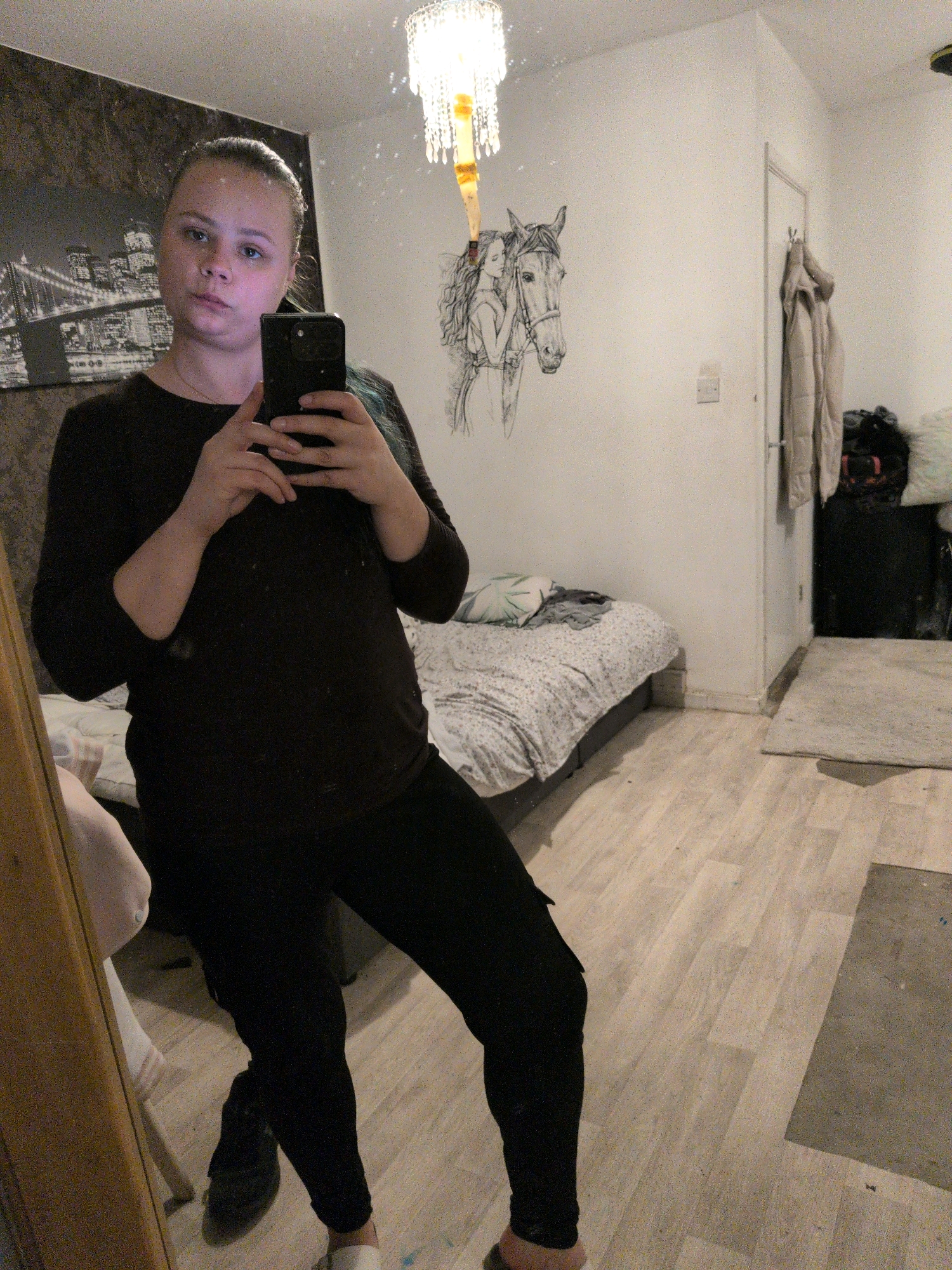A young woman with blonde hair pulled back stands in a bedroom and takes a mirror selfie. She is wearing a dark long-sleeved shirt and black pants. The room has a bed with a patterned comforter, a drawing of a woman and a horse on the wall, and a door with coats hanging on it. The floor is made of light-colored laminate. There is a large mirror to the left of the woman, reflecting her image and a portion of the room. The lighting in the room appears to be from overhead, with a chandelier visible. The overall impression is of a casual, lived-in bedroom. The location in the city of Oxford, United Kingdom is not discernible from the image.