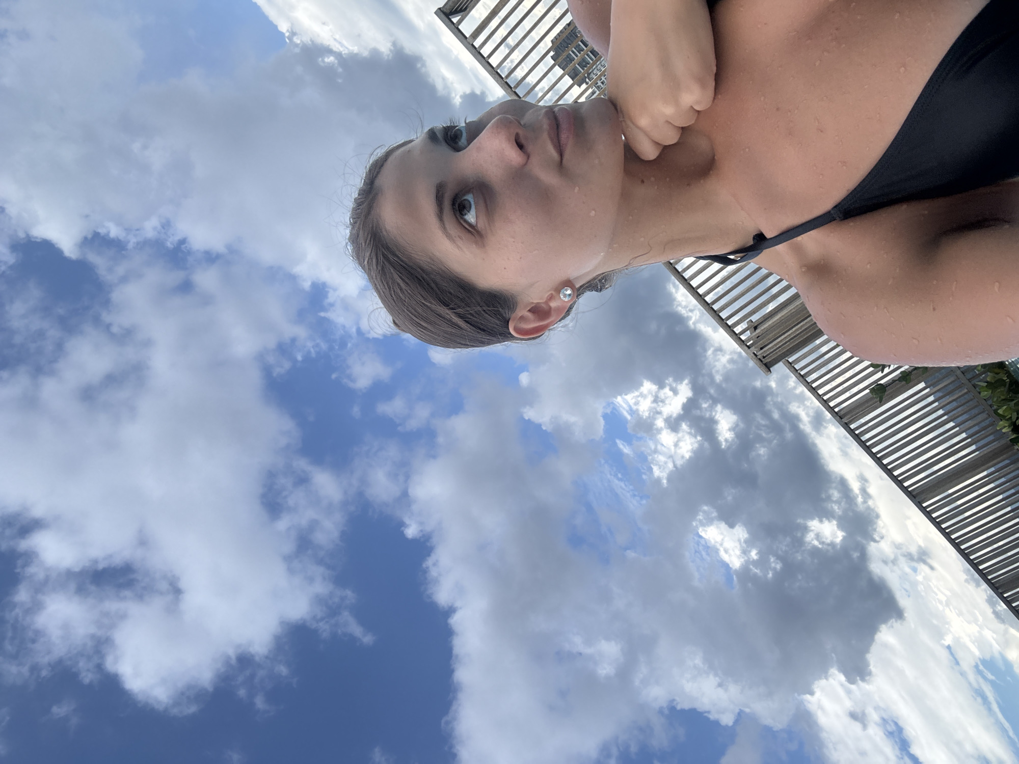 A woman wearing a black bikini is outside under a partially shaded structure with a view of the sky and clouds. She appears to be damp or wet, with visible water droplets on her skin. Based on the presence of skyscrapers visible through the shaded structure, the location is likely in Kuala Lumpur, Malaysia. The woman is holding her hand to her neck.