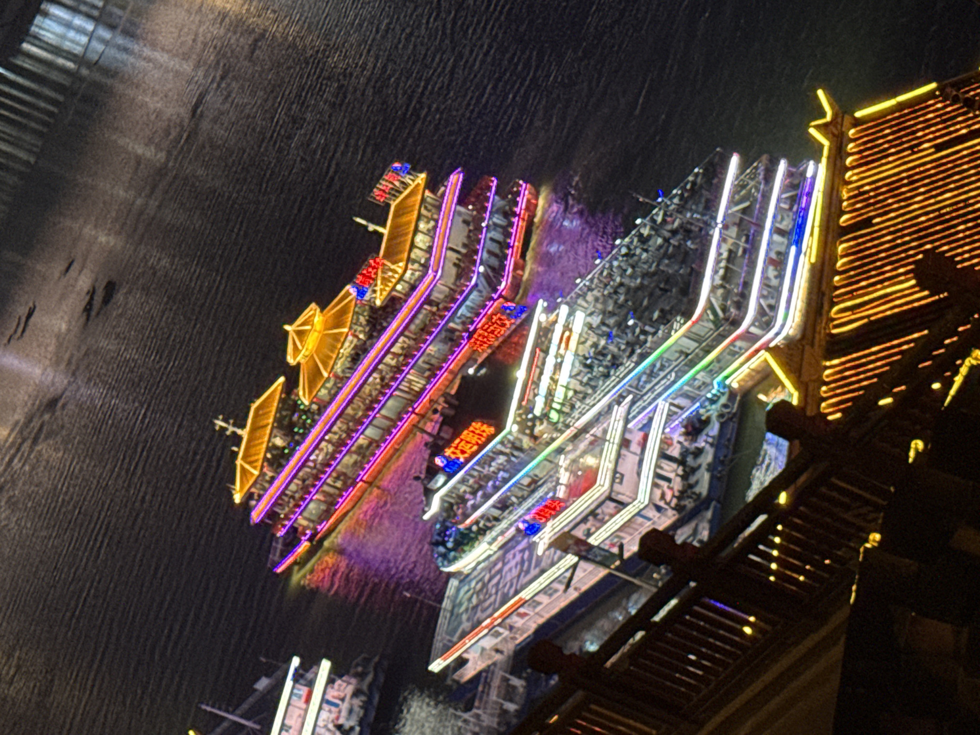 The image shows a night scene of illuminated tour boats docked on a body of water, likely the Yangtze River in Yuzhong District, China. The larger boat in the foreground, illuminated with pink and white lights, appears to be a multi-decked vessel with people visible on its upper levels. A smaller, similarly lit boat is docked alongside it. Further in the background, another boat, primarily lit with golden and purple lights, also appears to be docked. The water reflects the vibrant lights of the boats, creating a colorful display. A dark, elevated structure, possibly a bridge or promenade with railings and yellow lights, is visible in the lower left of the frame, suggesting a vantage point from which the photograph was taken. The overall impression is one of a lively waterfront at night.