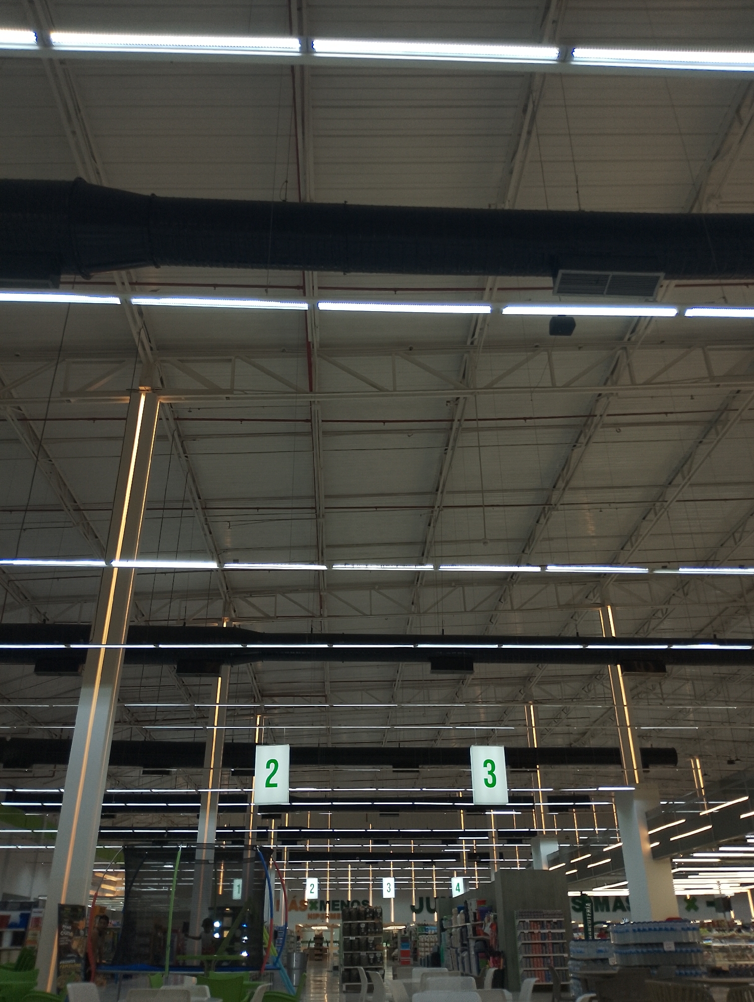 The image shows the interior of a large retail store, possibly a supermarket, in Porlamar, Venezuela. The high ceiling is a grid-like structure with fluorescent lighting fixtures running parallel. A large, black air duct is suspended from the ceiling. Below, hanging aisle markers labeled "2" and "3" are visible, guiding shoppers. Shelves stocked with merchandise are arranged in rows. Customers are partially visible in the background. Some objects such as green chairs, children's trampolines are present in front of the aisles.