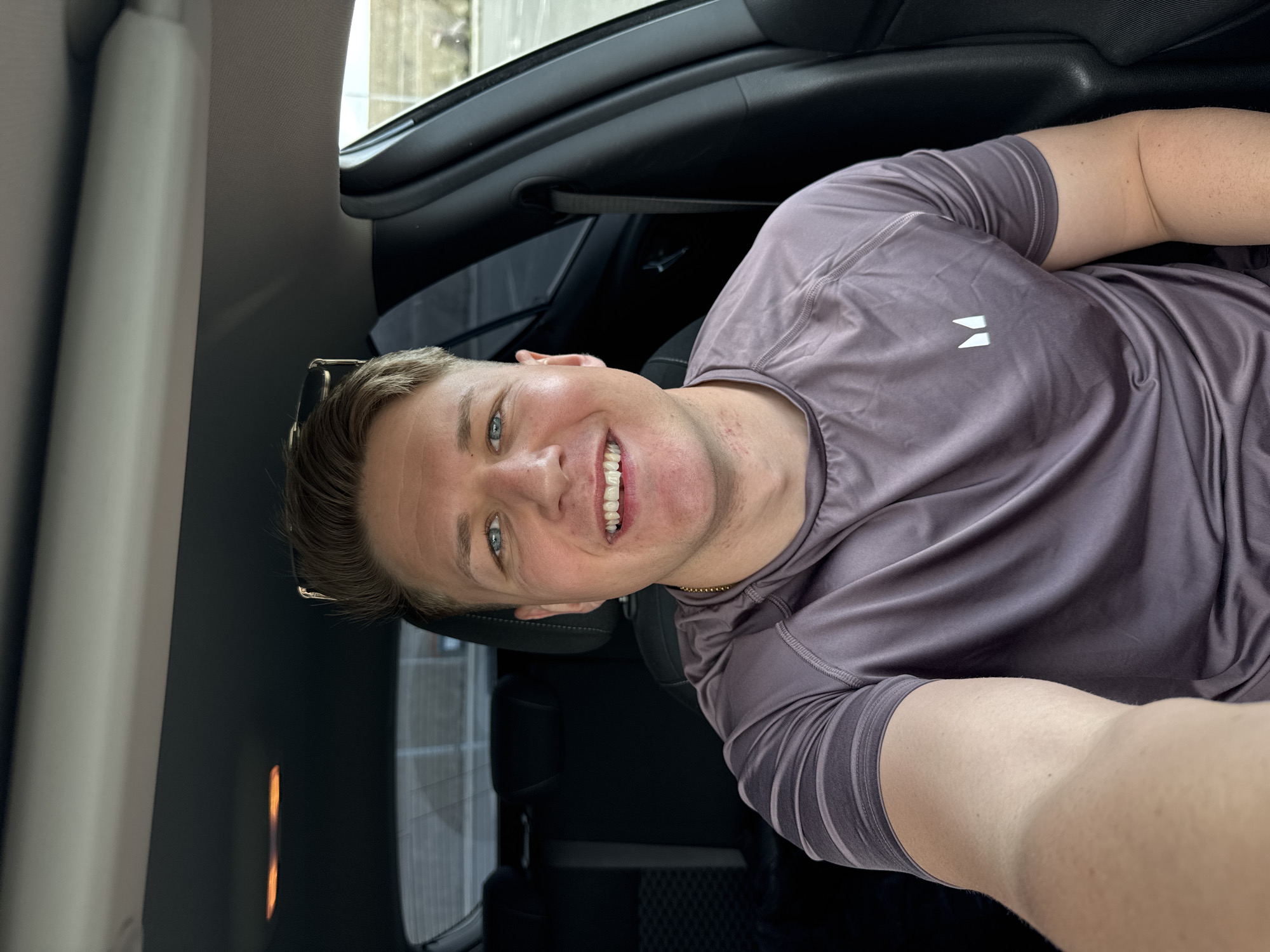 A man in a mauve athletic shirt is in the front seat of a car. He is smiling and taking a selfie. The background shows the car's interior and a blurry outdoor scene. It is impossible to confirm the location in Burgas, Bulgaria, without additional information.
