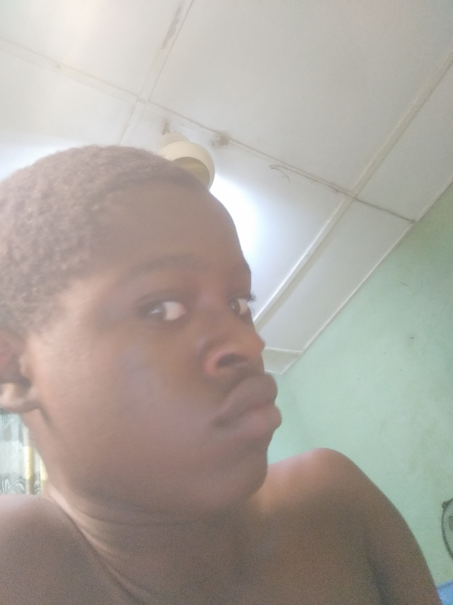 The image is a close-up selfie of a person with dark skin, short hair, and a slightly pouty expression. The background shows a white ceiling with a ceiling fan and a light fixture. A light green wall is visible on the right side of the frame. The location appears to be indoors.