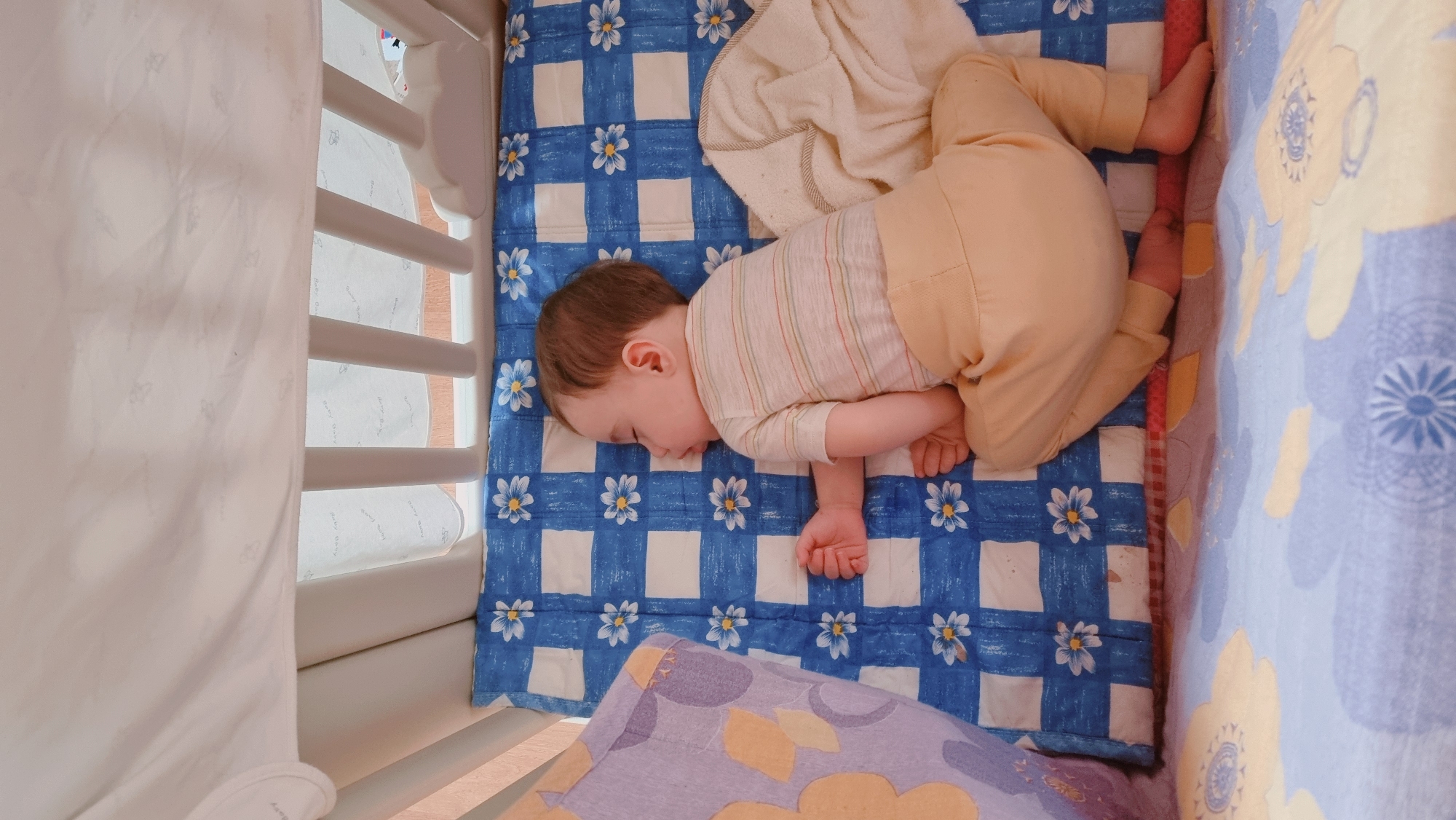 A baby sleeps on a blue-and-white quilt with a floral pattern in a white crib. The baby is wearing a striped shirt and light-colored pants. A tan-colored blanket is partially covering the baby. A patterned pillow is visible at the bottom of the frame. The location is indicated to be Dunnae-myeon, South Korea.