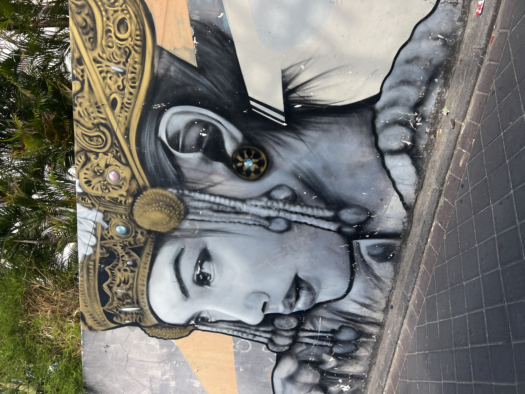 This is a photograph of a mural on a wall in Kuta, Indonesia, depicting the face of a woman adorned with a traditional headdress and jewelry. The mural is executed in shades of grey and black, with highlights of gold and blue on the headdress and earrings. The woman's face is rendered in profile, with her eyes looking to the left. She has dark hair styled in braids with decorative elements. The headdress is elaborate, featuring intricate golden patterns and embedded blue and pink jewels. Her earrings are circular and golden, with a blue stone at the center. The wall itself is textured and shows signs of wear, with peeling paint and some graffiti in other areas. Behind the mural, lush green foliage, likely palm trees, is visible, suggesting an outdoor setting. In the foreground, a paved walkway or sidewalk is depicted, made of dark square tiles with a border of reddish-brown bricks. The overall scene suggests a public space, likely a street or alleyway, in a tropical urban environment like Kuta.