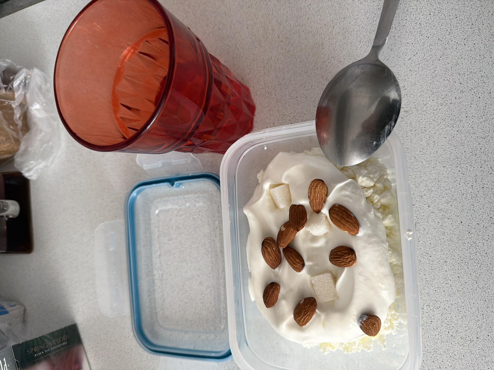 The scene is an overhead shot of a countertop in Leningradskaya, Russia. Visible are a red plastic cup lying on its side, a plastic container with a blue rim containing cottage cheese topped with yogurt, almonds, and candied fruit, a spoon resting on the cottage cheese, an open, empty plastic container with a blue rim, and a black tea box labeled "Spring Melody". The countertop has a speckled white and gray pattern. The lighting is bright and even, suggesting indoor light.