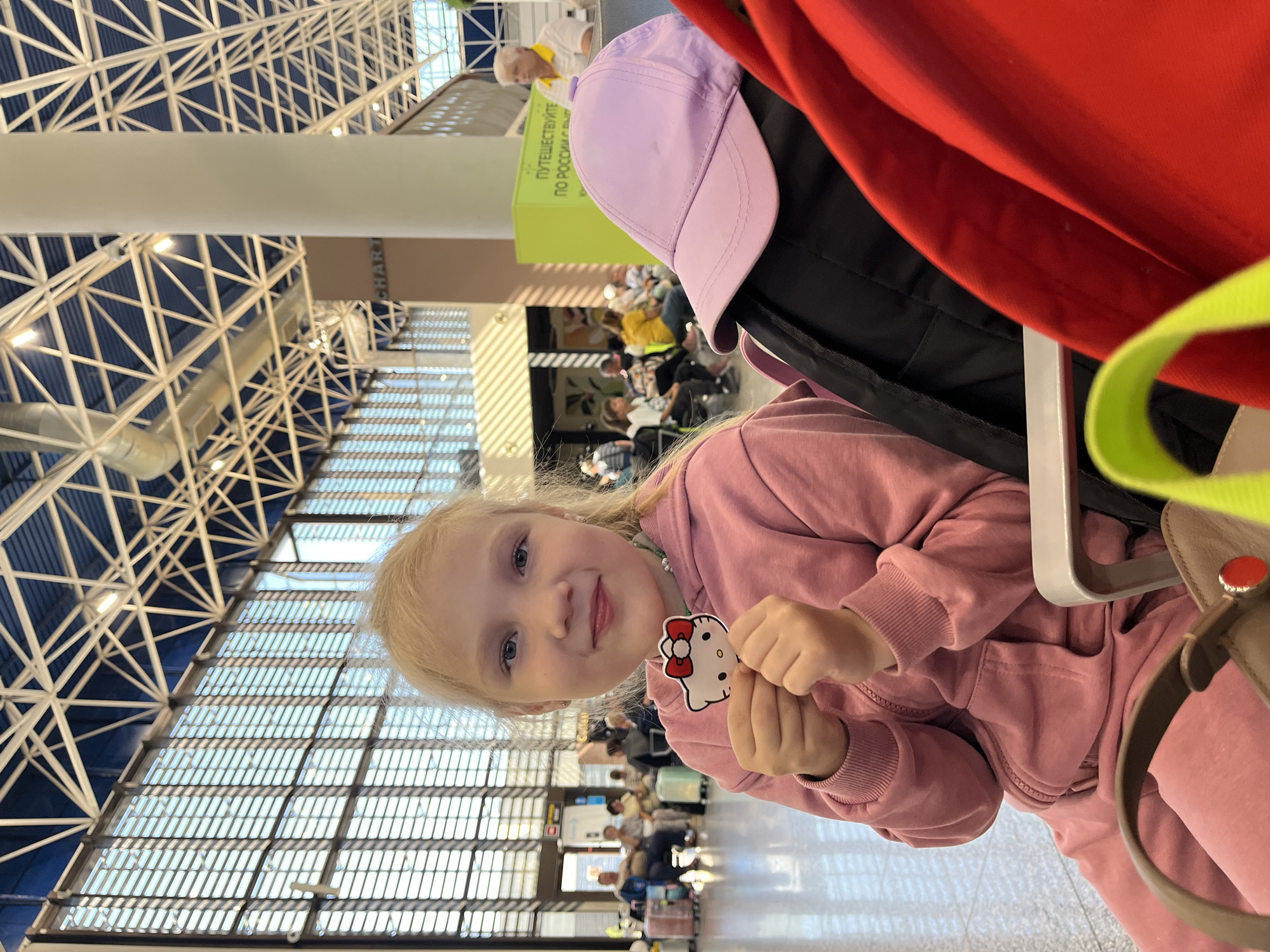 A young blonde girl with blue eyes, wearing a pink tracksuit, sits in an airport waiting area and holds up a Hello Kitty sticker. She is looking at the camera and smiling. In the background, other people are seated, waiting. Luggage can be seen in the foreground and background. The environment appears to be a modern airport terminal with large windows and a high, exposed ceiling structure. The signage in the background includes text in Russian, indicating the location is Sochi, Russia. A purple baseball cap and a red bag are visible in the foreground, partially obscuring the girl.