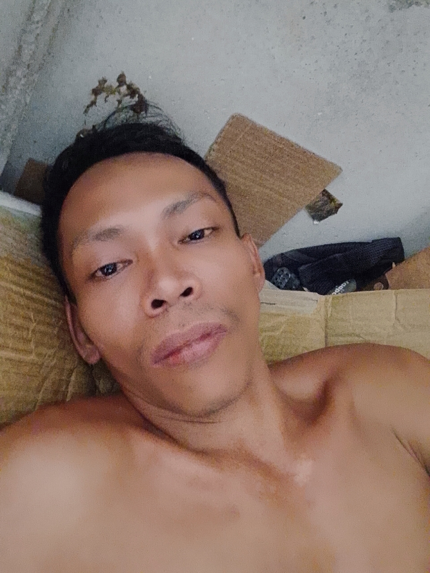 The image shows a shirtless man lying down, looking at the camera. He appears to be indoors. Beside the man are pieces of cardboard and what appears to be a black bag. The location is Bukit Ketok, Indonesia.