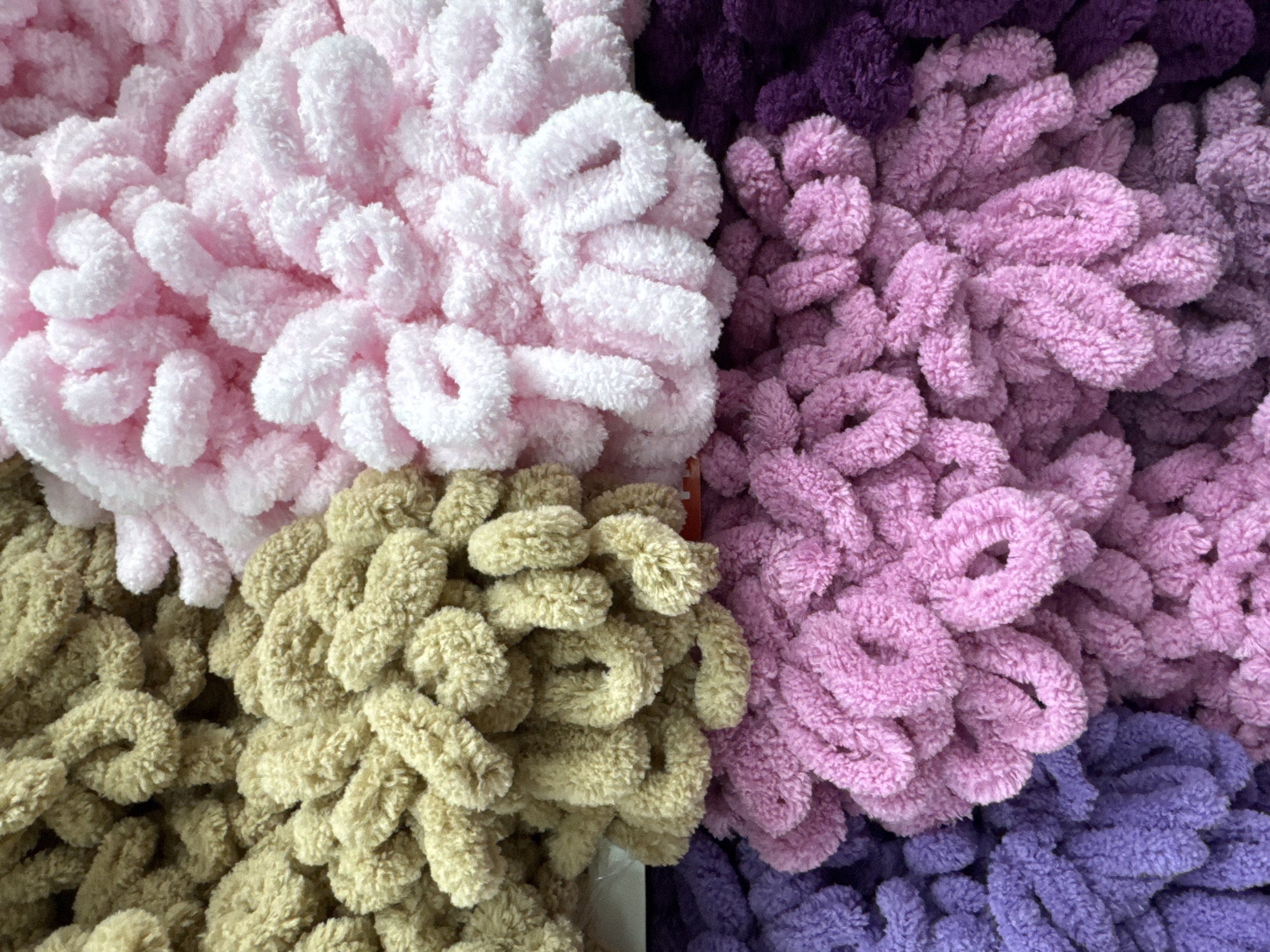 This close-up, overhead photograph, taken in Saint Petersburg, Russia, captures a vibrant display of four different colors of fluffy, looped yarn. The image is divided into four main sections by the arrangement of the yarn.
In the top-left quadrant, there is a pile of very light pink, almost off-white, looped yarn with a soft, plush texture. Adjacent to it, in the top-right section, is a mass of medium pink-purple yarn, which transitions into a darker purple in the upper right corner. The bottom-left quadrant features a cluster of muted yellow, beige, or olive-green looped yarn. Completing the arrangement, the bottom-right section displays predominantly darker purple looped yarn, with hints of the medium pink-purple bordering it, suggesting a gradient or mixed collection.
The yarn appears to be "loop yarn" or "finger knitting yarn," characterized by its pre-formed loops and soft, chenille-like texture. No people or specific activities are depicted, indicating the photo's focus is solely on the colorful yarn selection. The lighting suggests an indoor setting, possibly within a craft store or home. There is no visible text on any packaging or labels. The overall impression is one of tactile richness and a pleasing color palette.