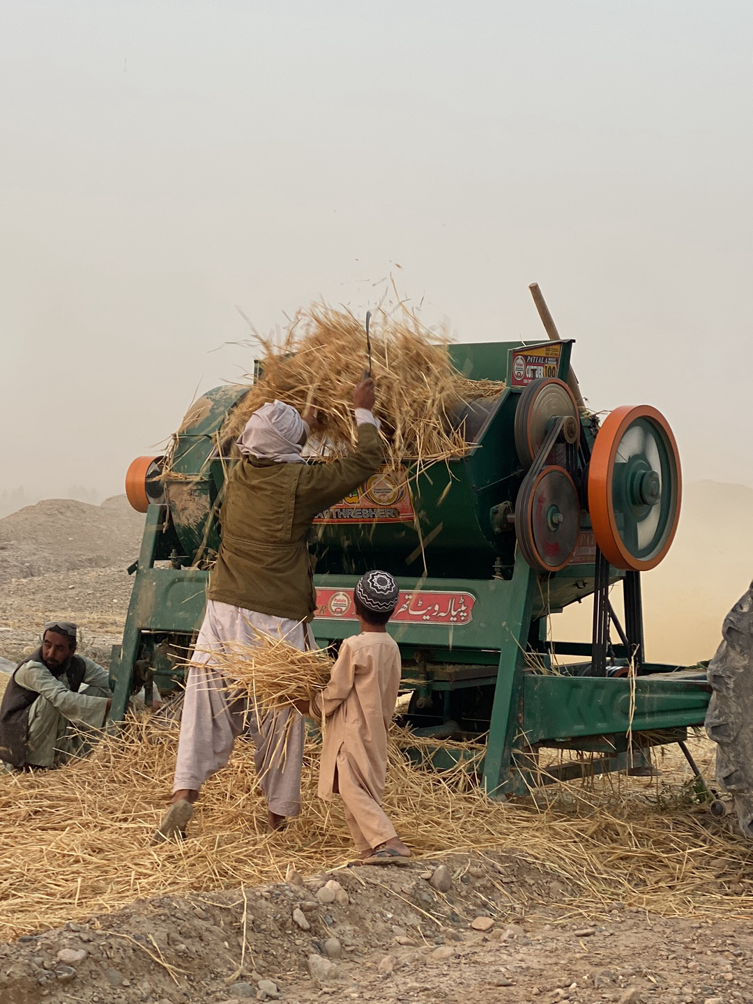 In Alaqadari Daman, Afghanistan, a group of people is engaged in agricultural work. A man wearing a jacket and loose pants is feeding sheaves of straw into a green threshing machine with orange wheels. A young boy in a light brown outfit stands beside him, also holding a bundle of straw. Another man sits on the ground to the left of the machine. The ground is covered in straw. The sky is hazy.