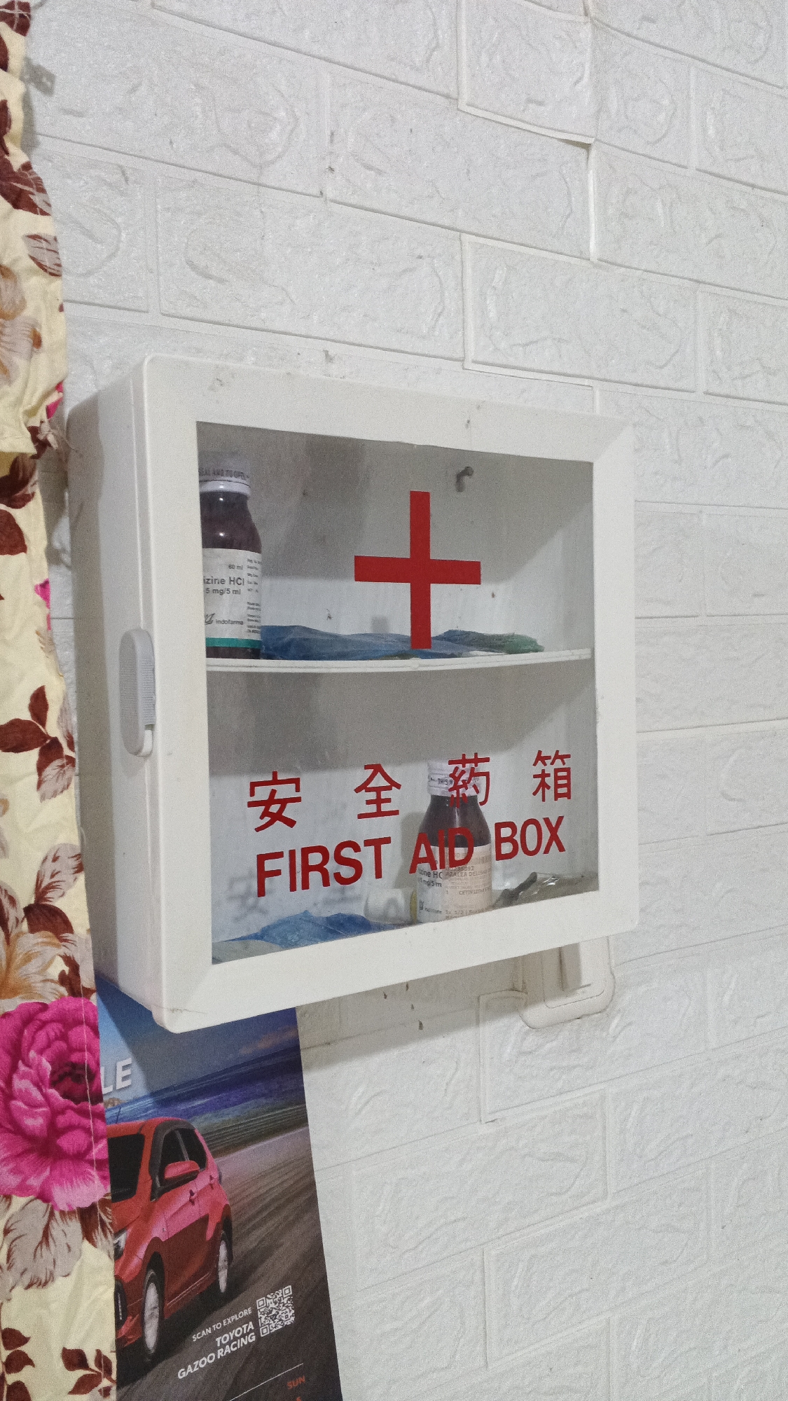 An indoor shot shows a white, wall-mounted first-aid box with a transparent door. Inside, there are two bottles of medicine, one on the top shelf and one on the bottom shelf, and a red cross symbol is affixed to the back wall of the box. Below the first-aid box, a portion of a poster with a red car and the text "GAZOO RACING" is visible. The wall behind the first-aid box is covered in white brick-patterned wallpaper. To the left of the first-aid box, a floral-patterned fabric is draped. The image was taken in Kampung Rempak, Indonesia.