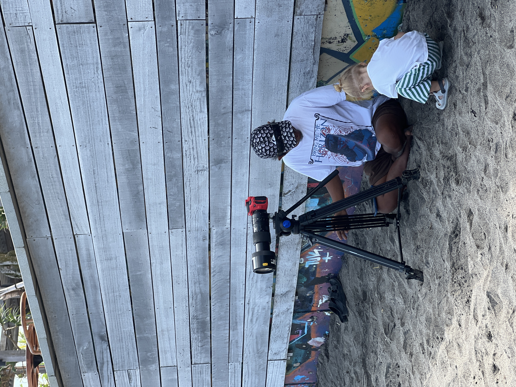 A person wearing a white t-shirt with a graphic print and a patterned baseball cap sits on dark sand in front of a weathered grey wooden wall. The wall has graffiti art at its base. A professional camera with a long lens is mounted on a tripod in front of the person. To the right of the person, a young child wearing a white t-shirt and green and white striped pants kneels on the sand, facing away from the camera. Black sandals are visible on the sand to the left of the tripod. The scene appears to be outdoors, possibly near a beach or coastal area. The lighting suggests it is daytime. The location is identified as North Kuta, Indonesia.